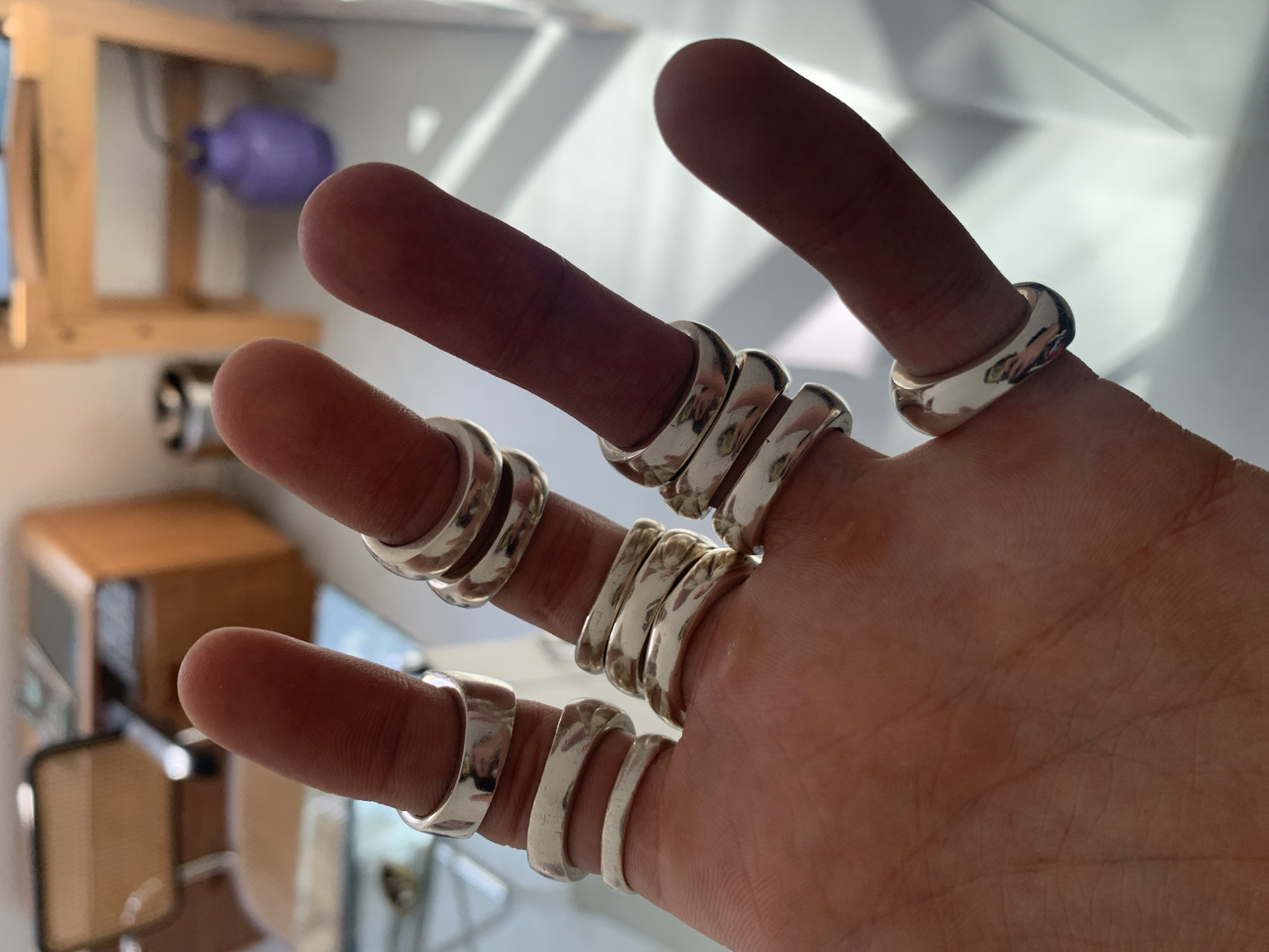 A close-up photograph shows a person's hand with multiple silver rings on each finger. The hand is positioned in the foreground with fingers splayed. Each finger except the thumb has two or three rings. The rings appear to be wide bands, likely made of polished metal.
The background is blurred but hints at an indoor environment with a wooden cabinet, a purple object hanging on a wall, and an interior space with bright light. The location in Buenos Aires, Argentina is not discernible from the visual information provided.