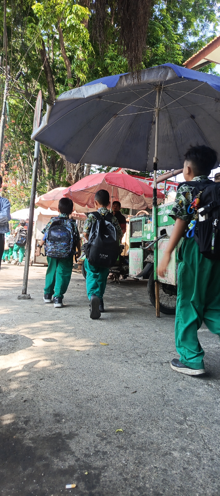 The image depicts three young boys walking away from the camera on a paved street in Krian, Indonesia. The boys are wearing matching green pants and patterned shirts, and each has a backpack on. The boy on the right has a black backpack with the word "FERAR" on it. The boy in the middle has a blue and black backpack with a detailed cityscape design. The boy on the left has a similar blue and black backpack.
They are walking under a large, dark blue umbrella, which is open and appears to be sheltering a food cart or stall. Behind the umbrella, another smaller, red umbrella is visible. The food cart is green and white, with a wheel on the right side. There are some white, round objects on display in the cart.
The environment appears to be outdoors, with trees and foliage visible in the background, suggesting a sunny day. The pavement is rough and textured, with some debris scattered on it, including a discarded cigarette butt in the lower right portion of the image. The overall scene suggests a street in a populated area, likely a commercial or school-adjacent zone.