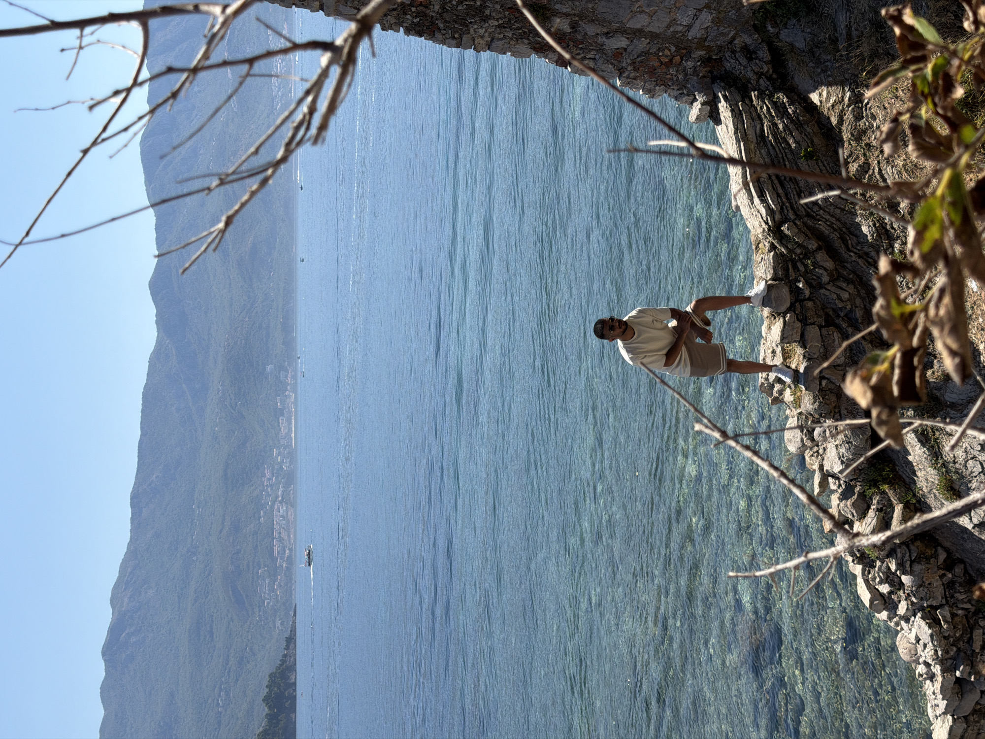 A young man wearing a light-colored t-shirt, khaki shorts, and white sneakers stands on a rocky outcrop overlooking the clear, blue water of the Adriatic Sea. He is looking to his left with sunglasses on. The backdrop features lush, green mountains that descend towards a coastal town with red-tiled roofs, suggesting the location is Budva, Montenegro. A boat is visible in the distance on the water. The foreground is partially obscured by dry, brown branches and leaves, and a crumbling stone wall adds to the ancient, rugged atmosphere of the scene. The sky is clear and bright blue.