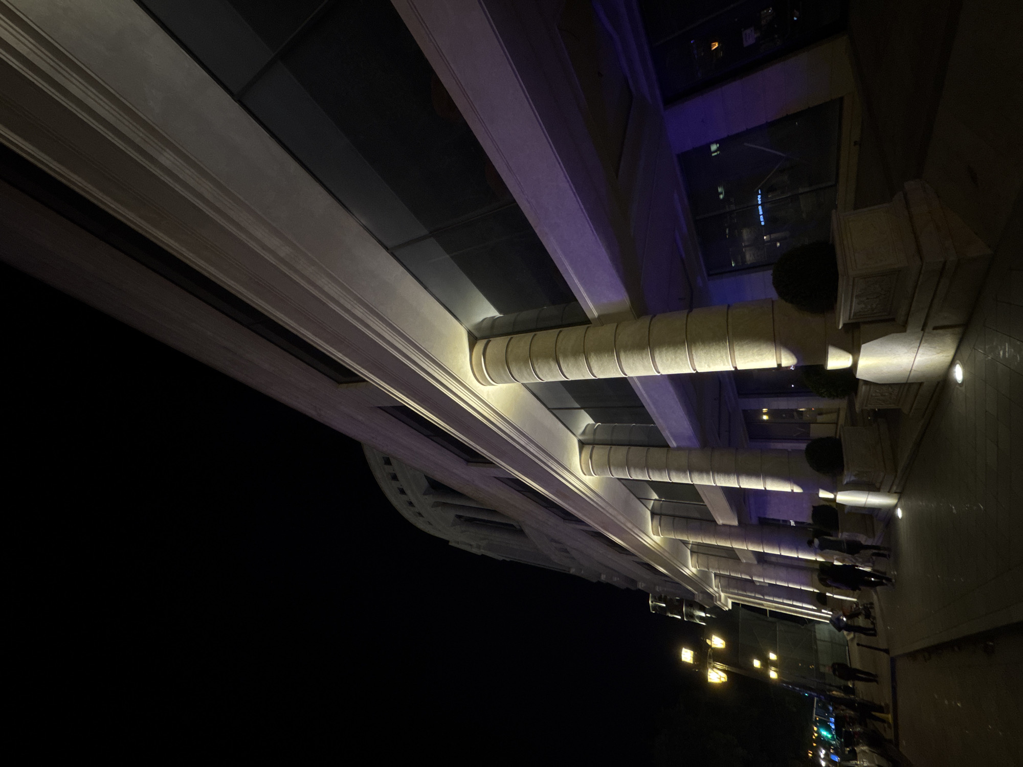 Captured at night, the image features an illuminated building in Yerevan, Armenia. The structure is characterized by a series of columns and windows. Beneath the building, a sidewalk is visible, occupied by several people. Streetlights cast a glow along the scene, enhancing visibility in the otherwise dark setting.