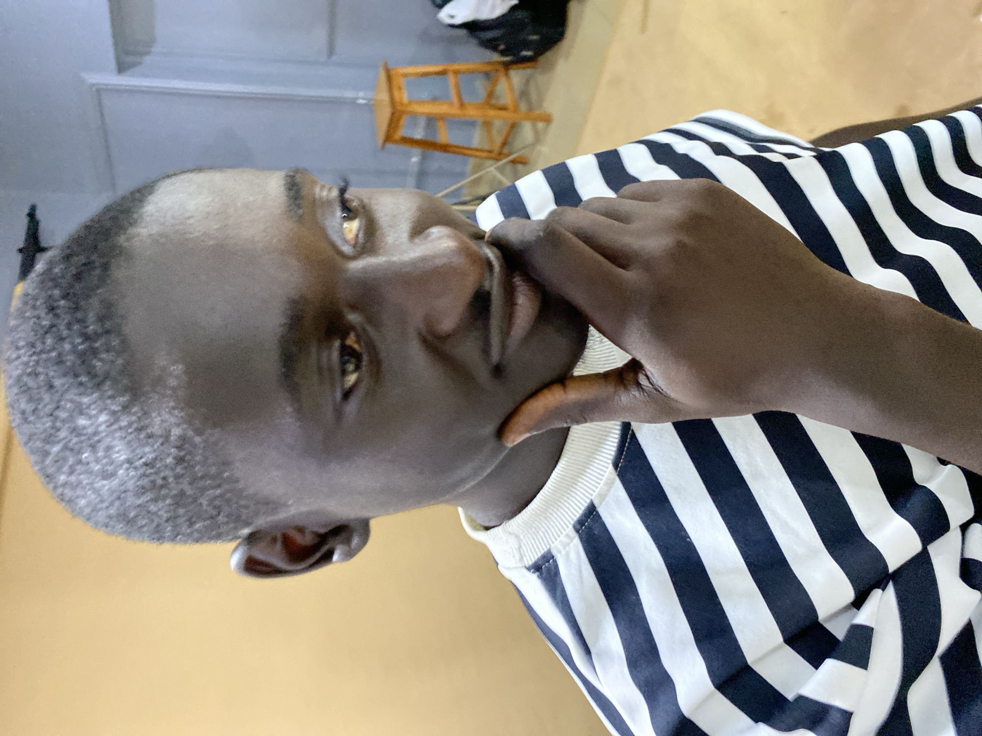 A dark-skinned man with close-cropped hair is holding his hand to his chin. He's wearing a striped black and white shirt. The location is Bukuru, Nigeria. A wooden stool and some clothing are in the background. The environment appears to be indoors.