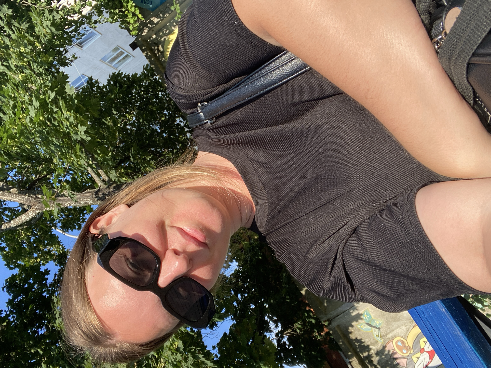 A young woman with blonde hair is taking a selfie outdoors on a sunny day, her face partially obscured by black sunglasses. She is wearing a black, ribbed short-sleeved shirt and has a black purse slung over her shoulder. The background features a large tree with green leaves, a building, and a blue bench. Graffiti is visible on a wall behind her. The scene appears to be in a public outdoor space.