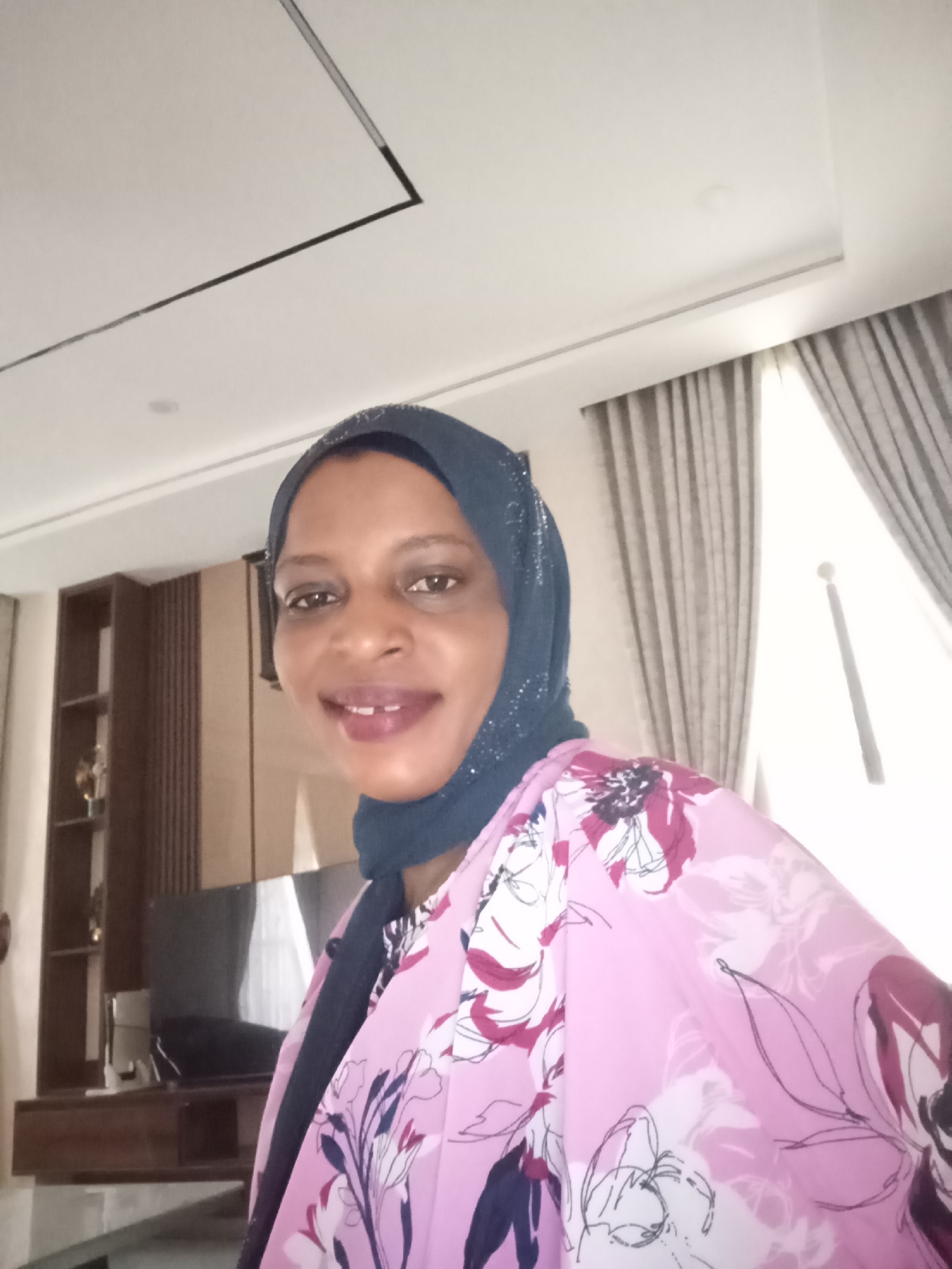 The image shows a woman wearing a dark blue hijab and a pink floral patterned top, possibly a kimono or robe. She is smiling slightly. Behind her, there is a built-in cabinet with open shelves and closed compartments. A dark-colored television is also visible on the cabinet. There is a window to the right with light grey curtains. The ceiling is white with recessed lighting. The location is Unguwar Rimi, Nigeria.