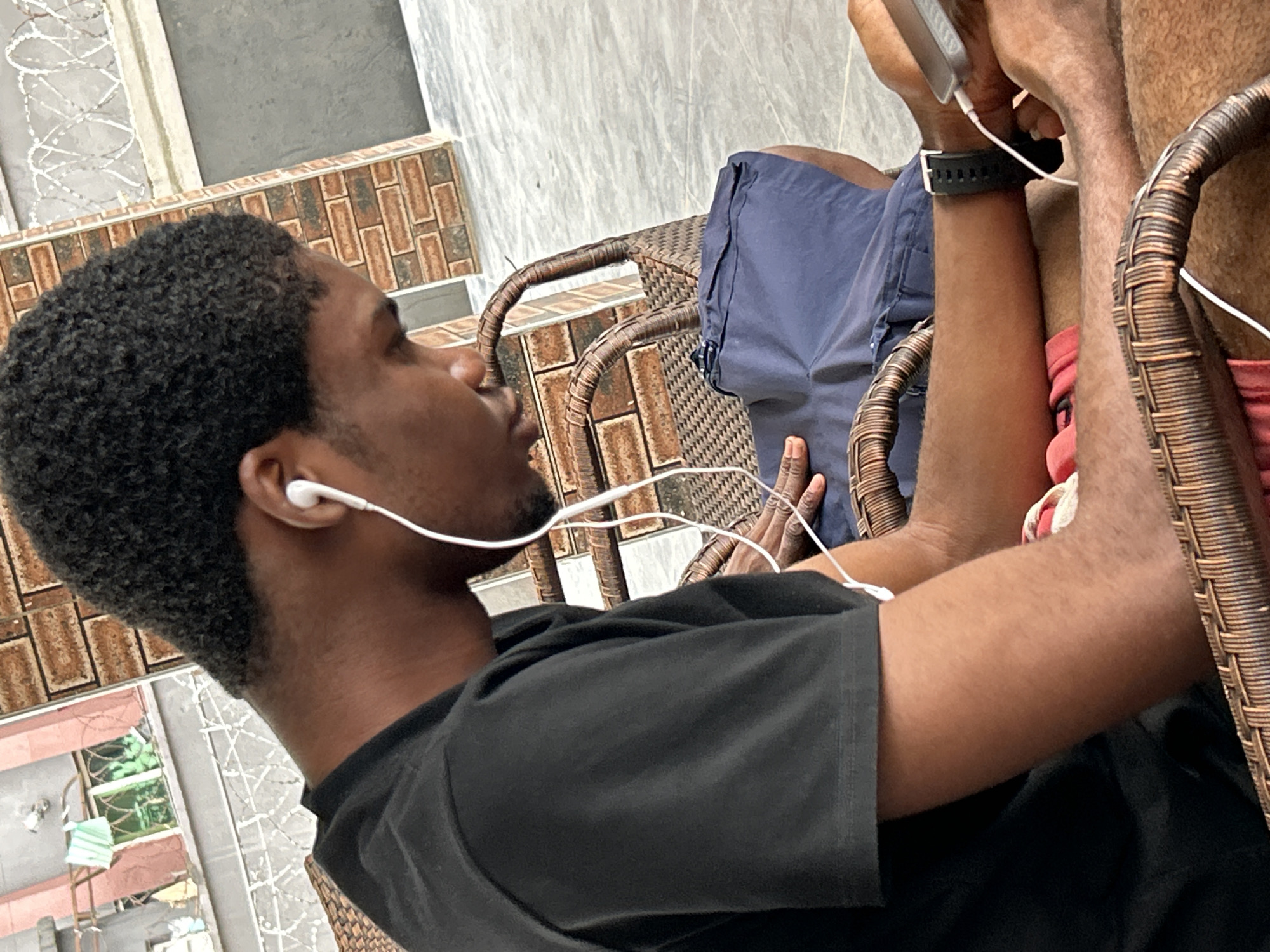A young Black male is seated on a wicker chair outdoors, facing to the right. He wears a black t-shirt and blue shorts. White earbuds are in his ears, with the cord extending out of frame. His right arm rests on his leg, and he is holding a smartphone in his hand. A black smartwatch is visible on his left wrist. The background consists of a brick wall, a metal fence with barbed wire, and a tiled floor. The scene appears to be in Ihiagwa, Nigeria.