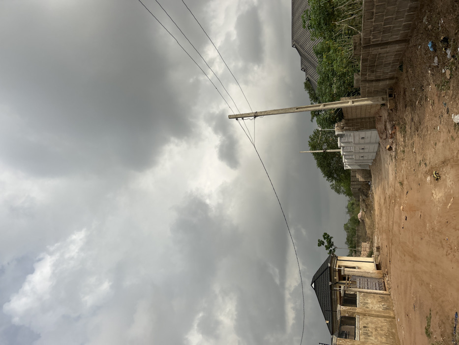 This image depicts a semi-developed area within Ilishan, Nigeria, under a heavily overcast sky. A reddish-brown dirt road, showing signs of vehicle tracks, occupies the foreground and extends into the midground. Litter, including plastic bags, is visible on the roadside.
To the left, a light-colored building with a dark, sloping roof is partially enclosed by a low, plastered wall with a light-colored metal gate. A single green tree stands behind this structure.
In the midground, towards the center-right, a concrete utility pole supports multiple overhead electrical wires that traverse the scene horizontally. Adjacent to the pole and extending to the right is a grey block perimeter wall, interspersed with newer, lighter grey pre-cast concrete fence panels. Beyond these walls, additional buildings with dark roofs are partially obscured by green vegetation, including small trees and shrubs. The background reveals undeveloped land with sparse greenery under the distant, hazy sky. No individuals are present in the scene.