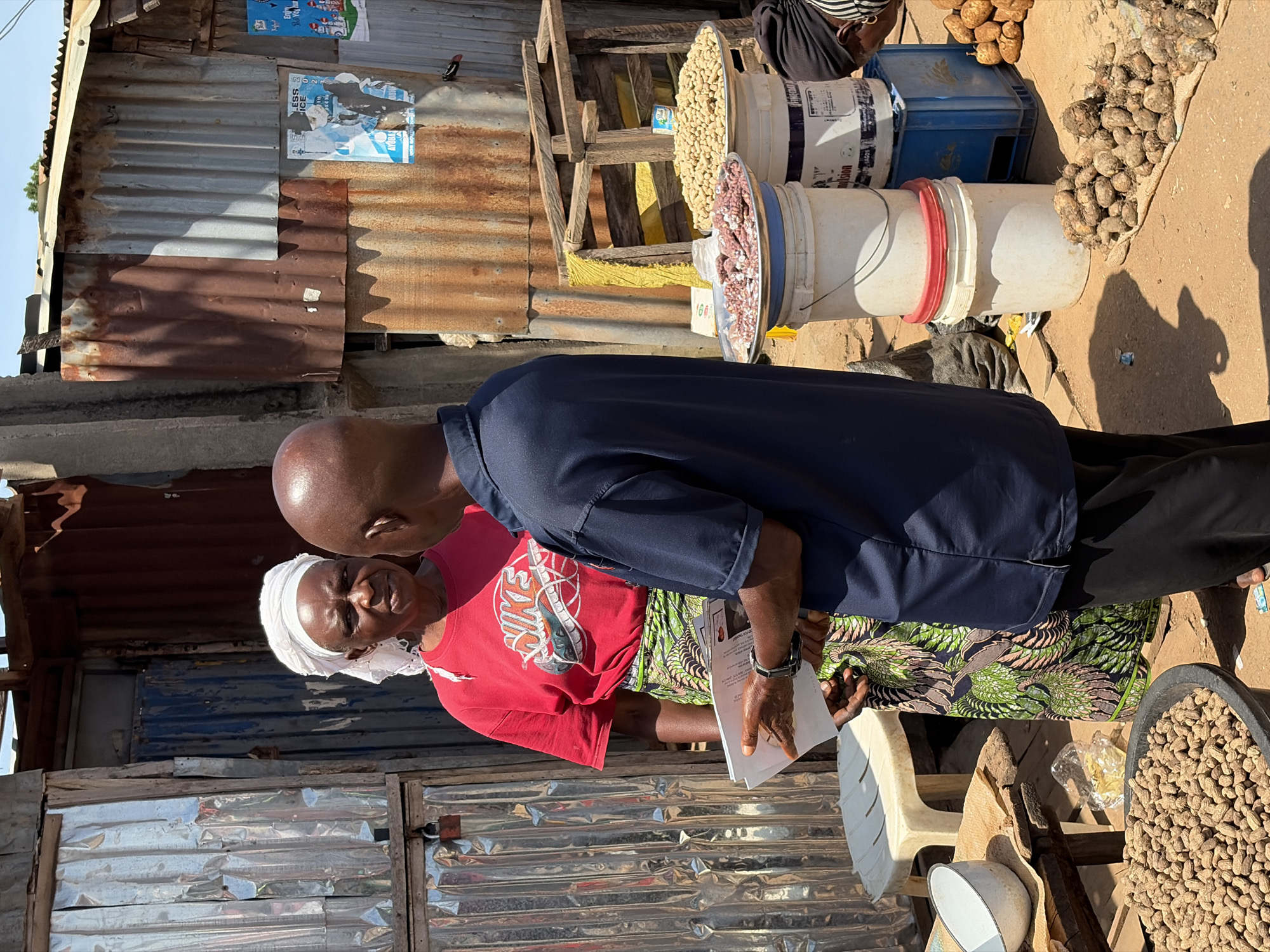 Two people, a woman and a man, are engaged in conversation in an outdoor market setting in Gura Top, Nigeria. The woman, with dark skin and wearing a white headscarf and a red t-shirt with a Nike logo, stands to the left, facing the man. She wears a vibrantly patterned green and black skirt. The man, with dark skin and a shaved head, is dressed in a dark blue short-sleeved polo shirt and black pants. He is positioned to the woman's right, facing her, and holds a few papers in his left hand, seemingly showing them to her.
The environment appears to be a makeshift market stall with corrugated metal walls and roofing, showing signs of rust and wear. To the right of the individuals, various goods are displayed on buckets and on the ground. Two large buckets are stacked, one white and one blue, with a third white bucket on top. On top of the buckets are two large platters of food; the front platter contains what appears to be dried meat or fish, while the rear platter holds a pile of peanuts. Another large bucket filled with peanuts is visible on the ground to the far left, and a pile of tubers or yams is spread on a mat on the ground to the right. A white plastic stool is also present near the woman. The ground is dirt, suggesting an unpaved market area. The sky is visible above, indicating it is daytime. The overall scene depicts a transaction or information exchange in a bustling, informal market environment.