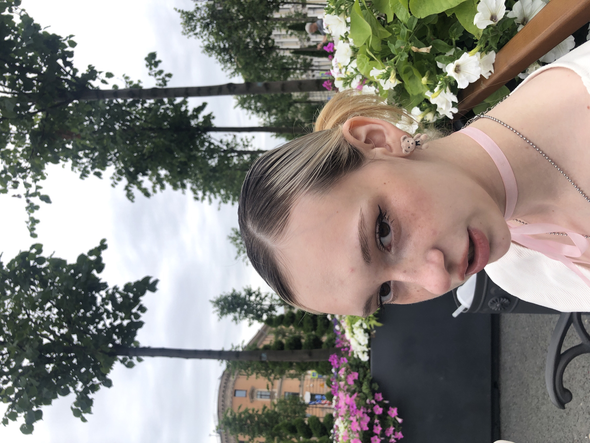 An outdoor, medium shot shows a young white woman with fair skin, freckles, and light brown hair pulled back into a ponytail. She is looking towards the right side of the frame. Her left ear is adorned with a teddy bear-shaped earring and she is wearing a light pink choker necklace with a silver chain. She has winged eyeliner and blush on her cheeks. She is wearing a white cardigan. In the background, there are trees, flowers, a building, and a cloudy sky. The scene appears to be in a city park or plaza.