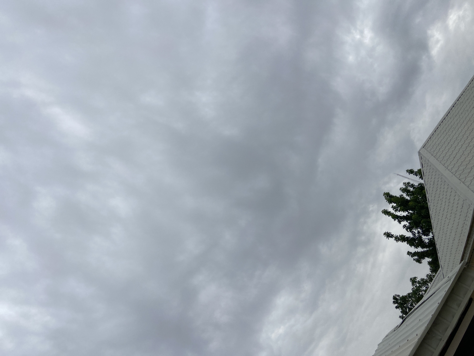 The image depicts a cloudy sky above a residential area. The foreground shows the white, tiled roofs of two houses, with one extending from the bottom left corner of the frame. The roofs are modern and appear to be in good condition. Behind the roofs, lush green trees with dense foliage are visible. The sky is overcast with a layer of grey clouds, suggesting a subdued or impending rainy weather. The scene appears to be a typical suburban or semi-urban landscape in Gura Top, Nigeria. No people or specific actions are depicted in the image.