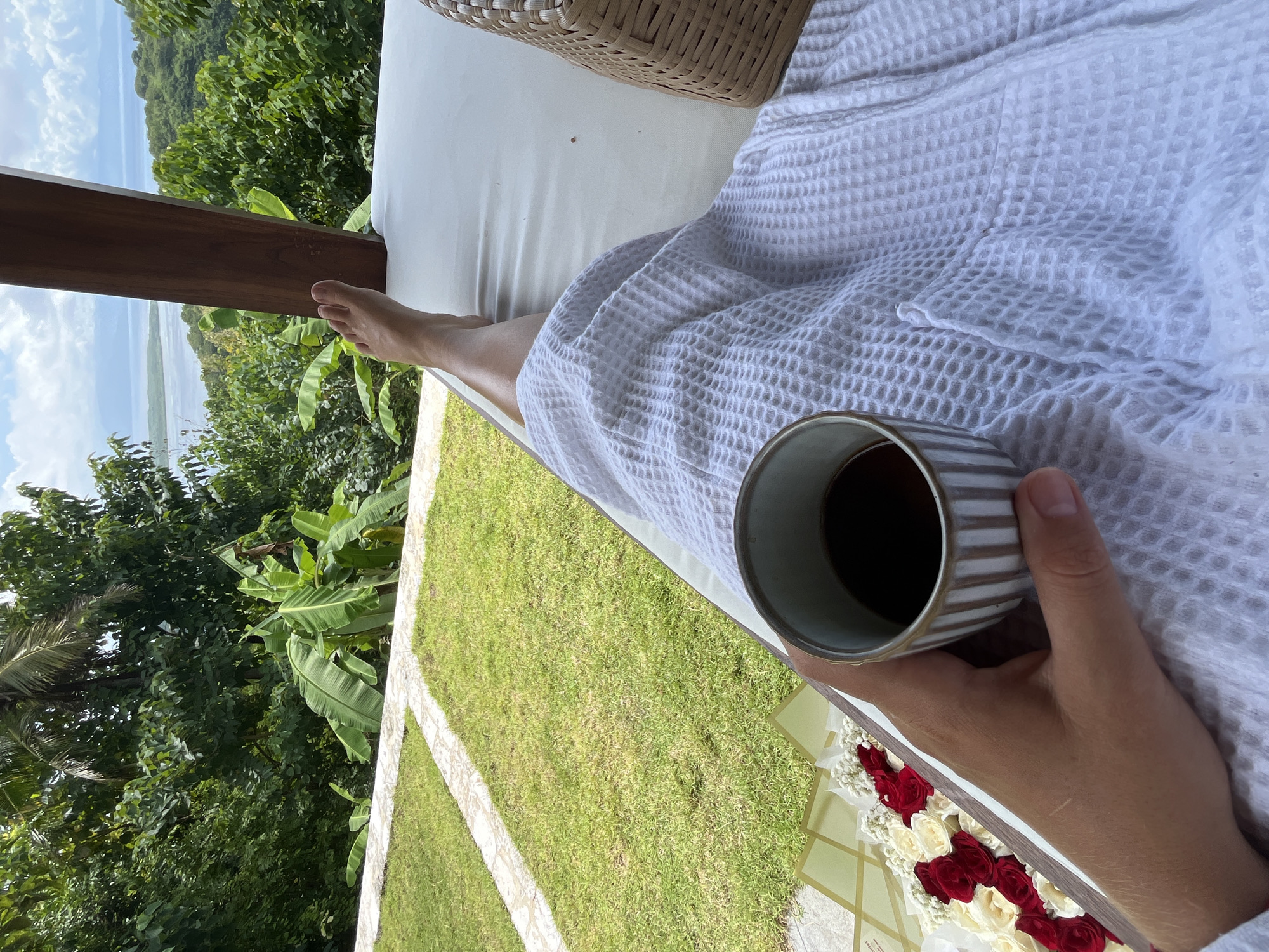 A person is reclining on a white cushioned chaise lounge, clad in a white waffle-weave robe, holding a striped mug filled with a dark liquid, presumably coffee, in their right hand. A bare foot rests on the lounge. Next to the person are red and white roses arranged in a row, resting on a pale tile surface. To the side is a manicured lawn, surrounded by lush green trees and banana leaves, creating a tropical setting. In the distance, a body of water can be seen under a blue sky. The photo appears to be taken in Nusa Penida, Indonesia, based on the foliage and landscape.
