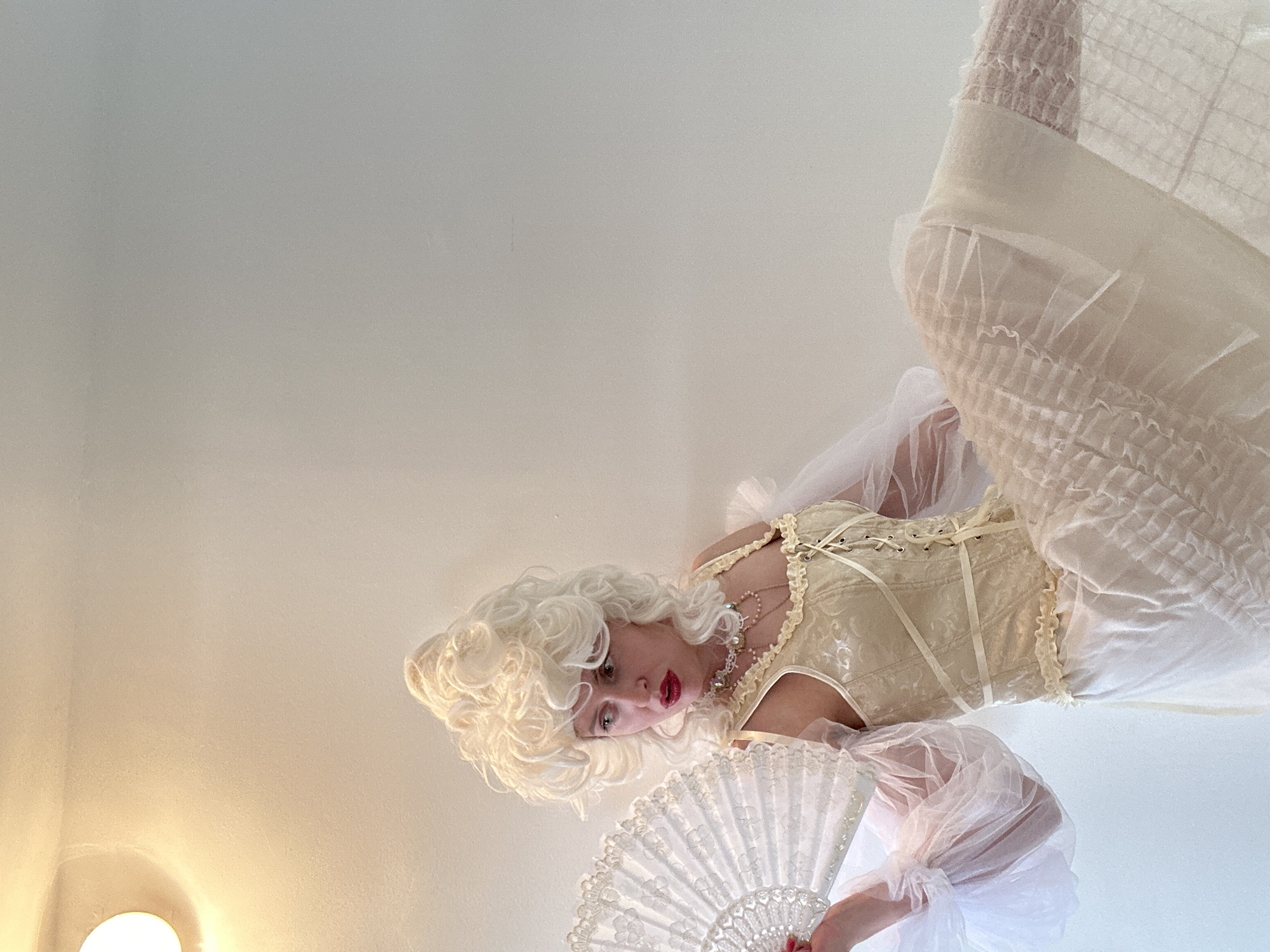 The image displays a person, appearing to be female, dressed in elaborate historical-inspired attire, captured from a low-angle perspective against a plain, light-colored background. The individual wears a voluminous, curly blonde wig, characteristic of 18th-century or Rococo fashion, and striking red lipstick. Her gaze is directed downwards and to the left, conveying an intense or theatrical expression.
Her costume includes a light-colored, possibly cream or champagne-toned, corset or bodice with intricate lacing down the front and delicate ruffled trim. She has sheer, puffy sleeves made of a white or cream fabric, resembling organza or tulle. A multi-strand necklace, appearing to be pearls with a central pendant, adorns her neck. In her right hand, she holds a white, ornately decorated folding fan with intricate patterns. Her lower garment is a full, light-colored skirt or petticoat, featuring ruffles and textured fabric details.
The background is a smooth, unadorned surface, likely a wall or ceiling, with a subtle color gradient transitioning from a warmer, yellowish tone at the bottom left to a cooler, off-white hue towards the top right. A circular light fixture emitting a warm glow is partially visible in the bottom left corner, indicating an indoor setting with artificial illumination. The scene suggests a posed photoshoot or a costumed portrayal, evoking a historical or fantasy aesthetic. No text is visible in the image.