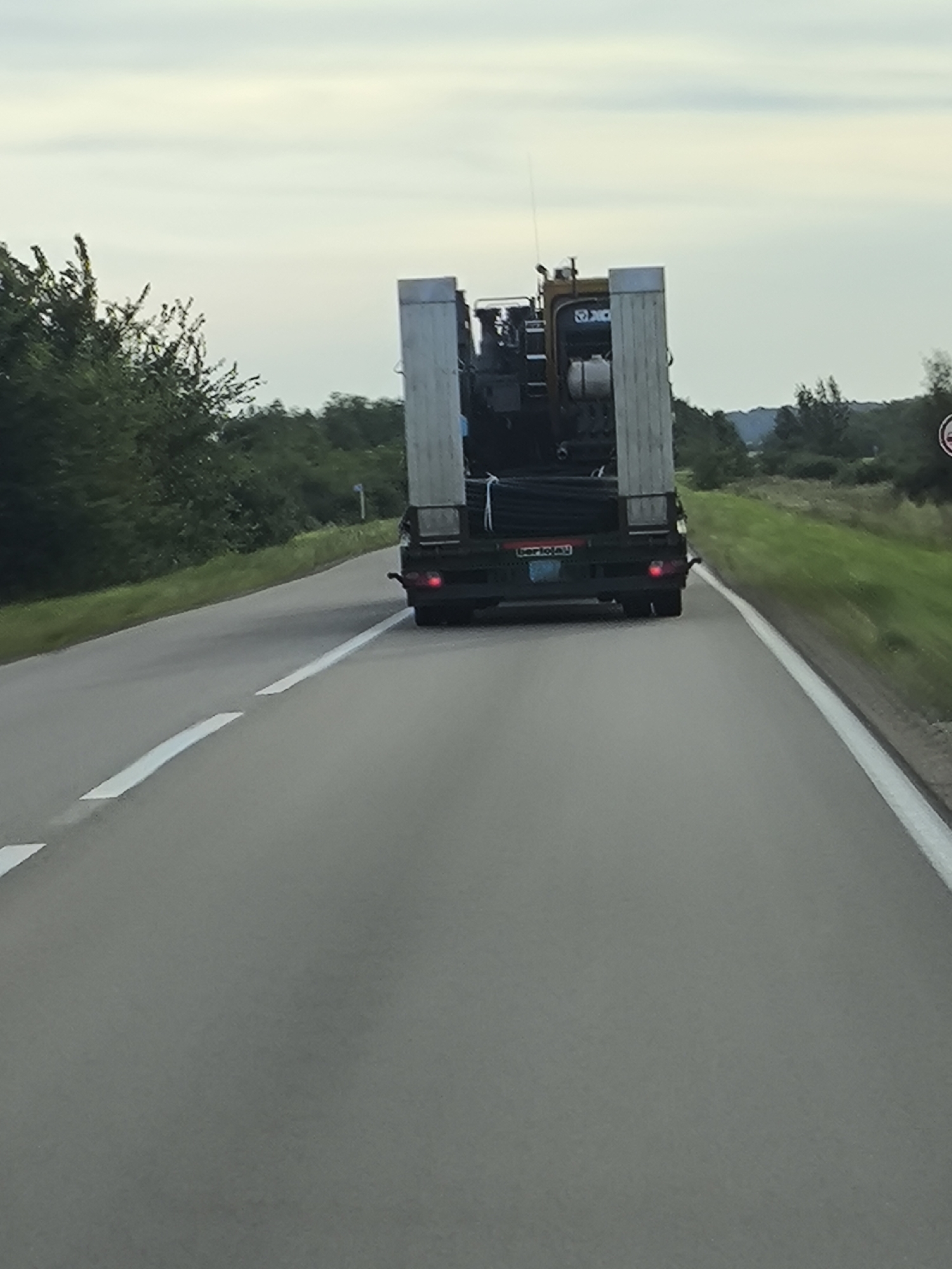 The image shows a rear view of a large truck driving on a paved road. The truck is carrying a load of what appears to be large pipes or cylindrical objects wrapped in black plastic. The truck itself is dark colored with some yellow and silver elements. The road has white dashed lines indicating lanes. On either side of the road, there is a grassy shoulder and then trees and vegetation. The sky is overcast. The location is identified as Станов ауылдық округі, Kazakhstan.