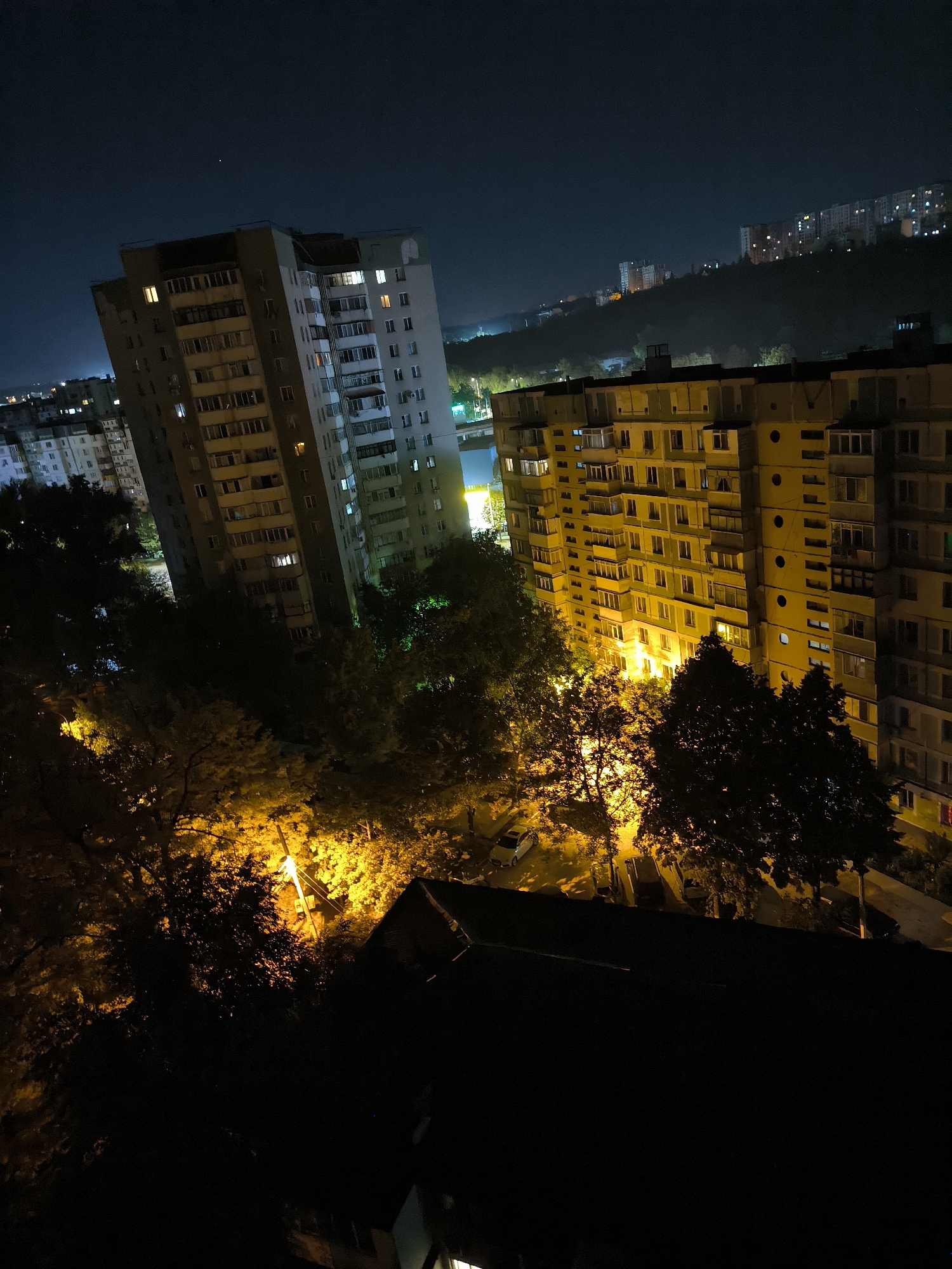 This image depicts an urban night scene in Chișinău, Moldova, focusing on apartment buildings and surrounding foliage illuminated by artificial lights. In the foreground, dark silhouettes of trees obscure the lower part of the frame, with a prominent street lamp casting a bright yellow glow on the foliage. Beyond the trees, two large, multi-story apartment buildings are visible. The building on the left is lighter in color and appears slightly tilted. Both buildings have numerous windows, many of which are illuminated, suggesting residential occupancy. Further in the background, a hillside is dotted with more apartment buildings, their lights creating a warm pattern against the dark landscape. The sky is dark and clear, with a single bright star visible in the upper center. The overall impression is one of a typical residential area at night, with the architecture characteristic of Eastern European cities.