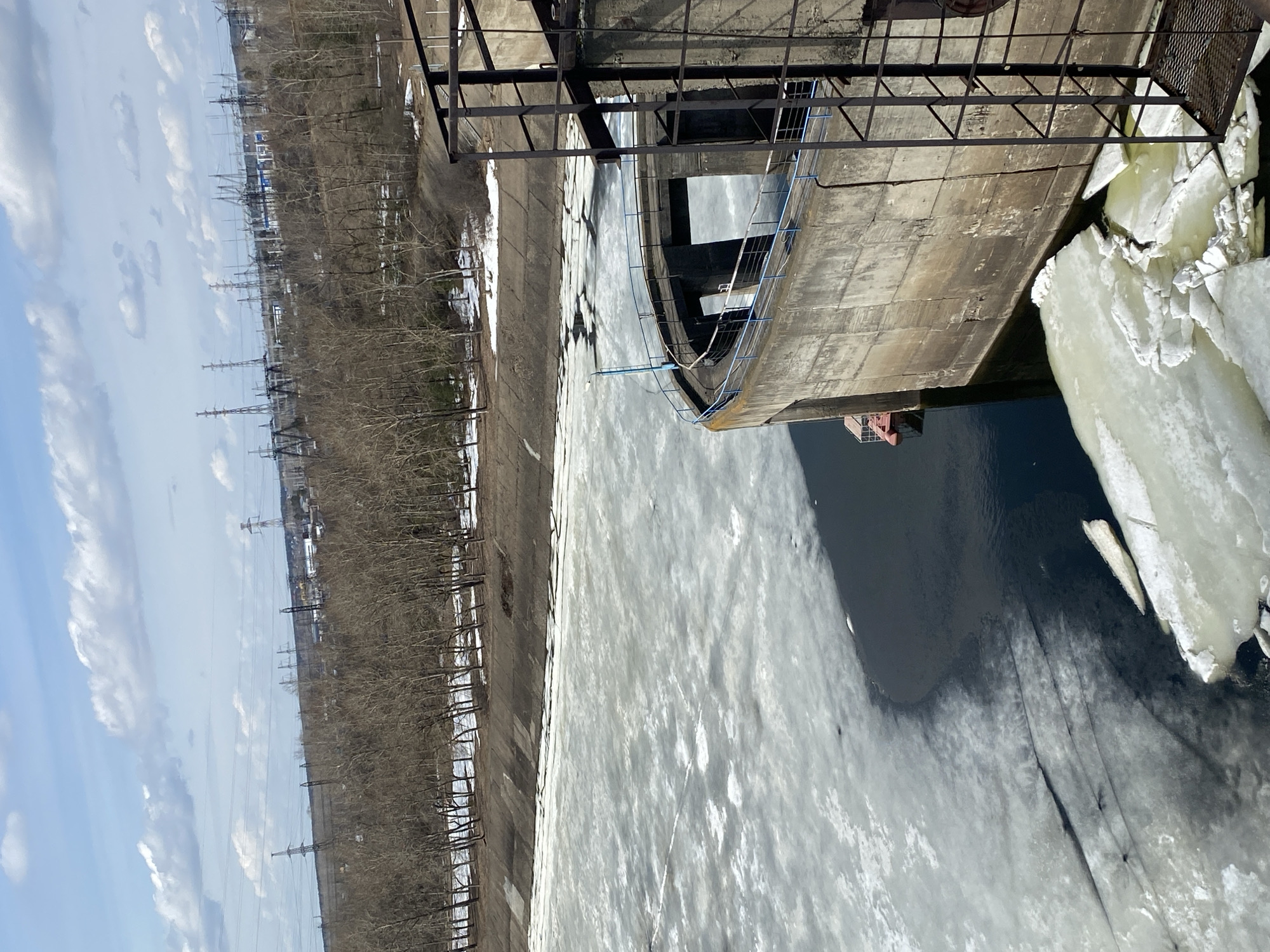 The image shows a section of the Kama River in Chaikovsky, Russia, during a period of ice breakup. A concrete structure, part of a dam or bridge support, protrudes over the partially frozen river. Large chunks of ice are clustered near the structure, with open water visible. Along the riverbank, there is a concrete embankment and a line of trees, behind which are power lines. The sky is partly cloudy. The scene suggests the transition from winter to spring.