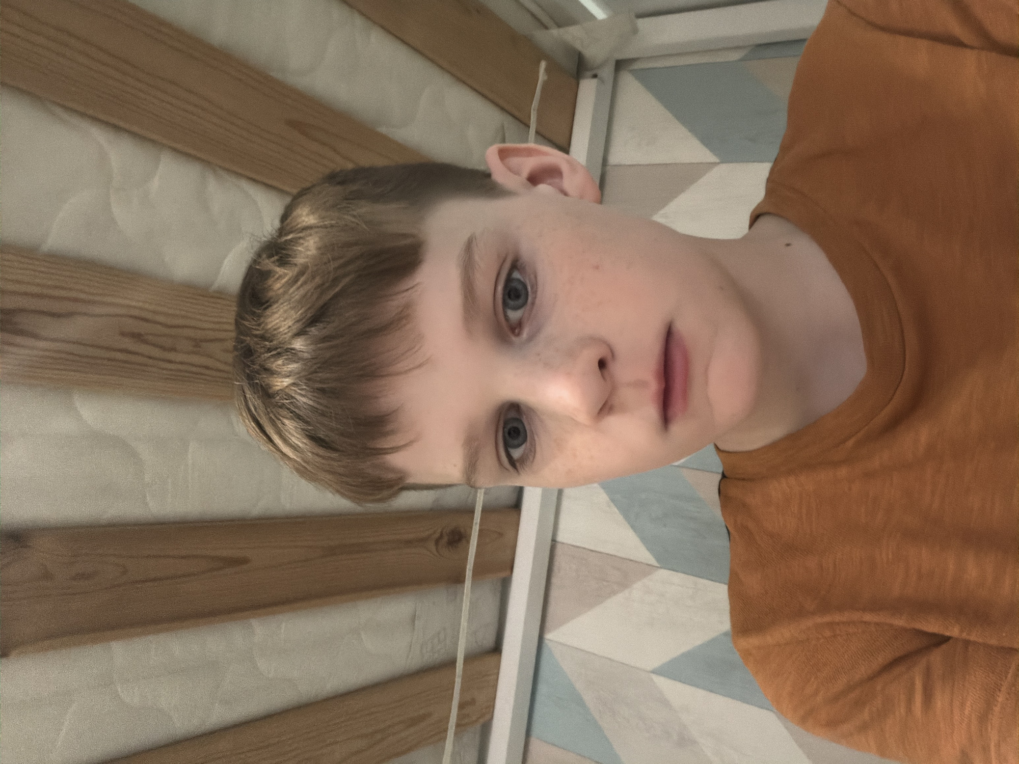 A close-up, slightly rotated view of a young person with fair skin, light brown hair, and blue eyes. The person is wearing an orange shirt. The background consists of a wooden-slat wall and a geometrically patterned wall with light blue and white colors. There is no indication of the location being in Zaprudnya, Russia.