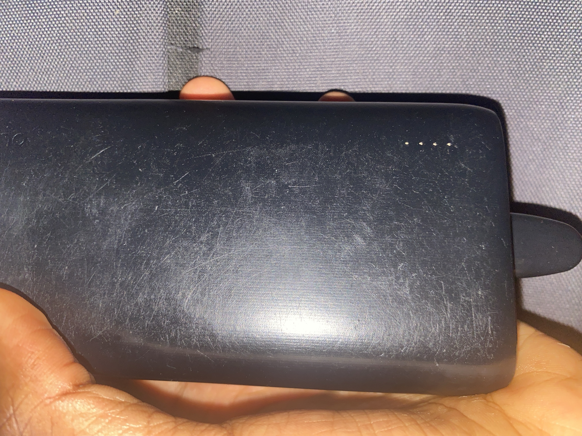 A person is holding a scratched, dark-colored power bank. The power bank has four indicator lights on the top edge and a rubberized tab on the right side. The background is a gray textured surface. The location is unknown, and there is no information to suggest that the location is in Gombe, Nigeria.