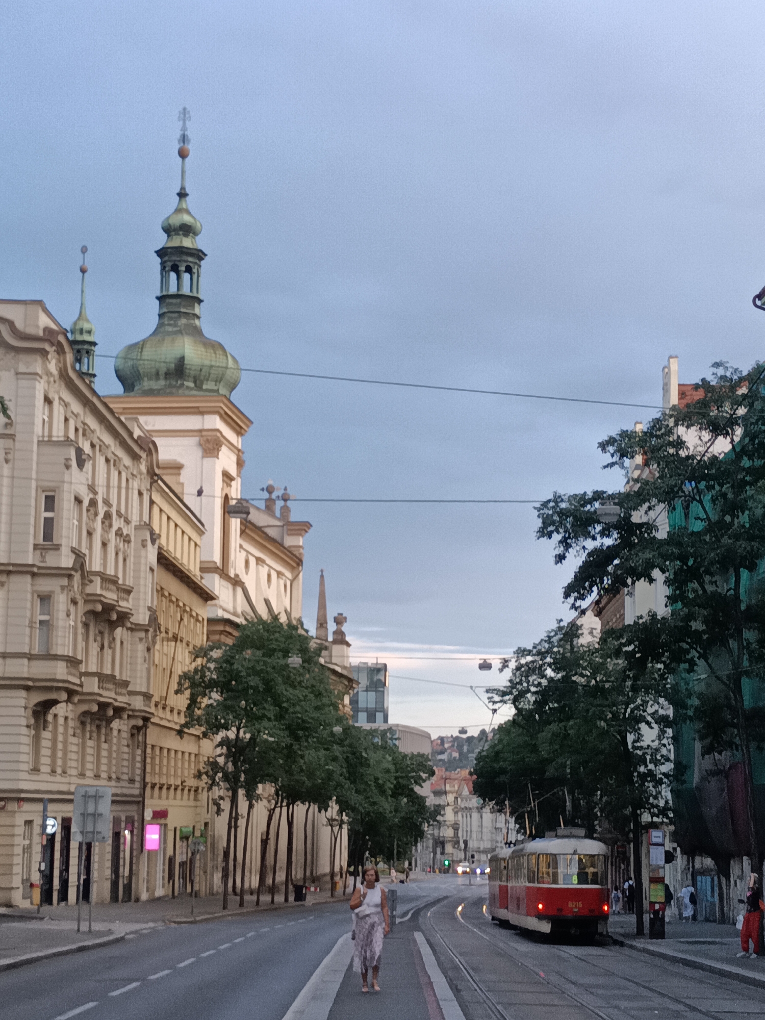 The image captures a street scene in Prague, Czechia, under a cloudy sky. A vintage red tram with the number 8215 is traveling on the tram tracks. Several buildings line the street, featuring architectural styles common in European cities, with intricate facades and balconies. Tall trees with green foliage are planted along the sidewalk, creating a sense of an avenue. In the foreground, a woman wearing a light-colored dress is walking away from the camera on the right side of the street. The street is paved, and tram wires are visible overhead. The overall impression is that of a bustling European city, likely during daylight hours, although the overcast sky suggests it might be late afternoon or early evening.