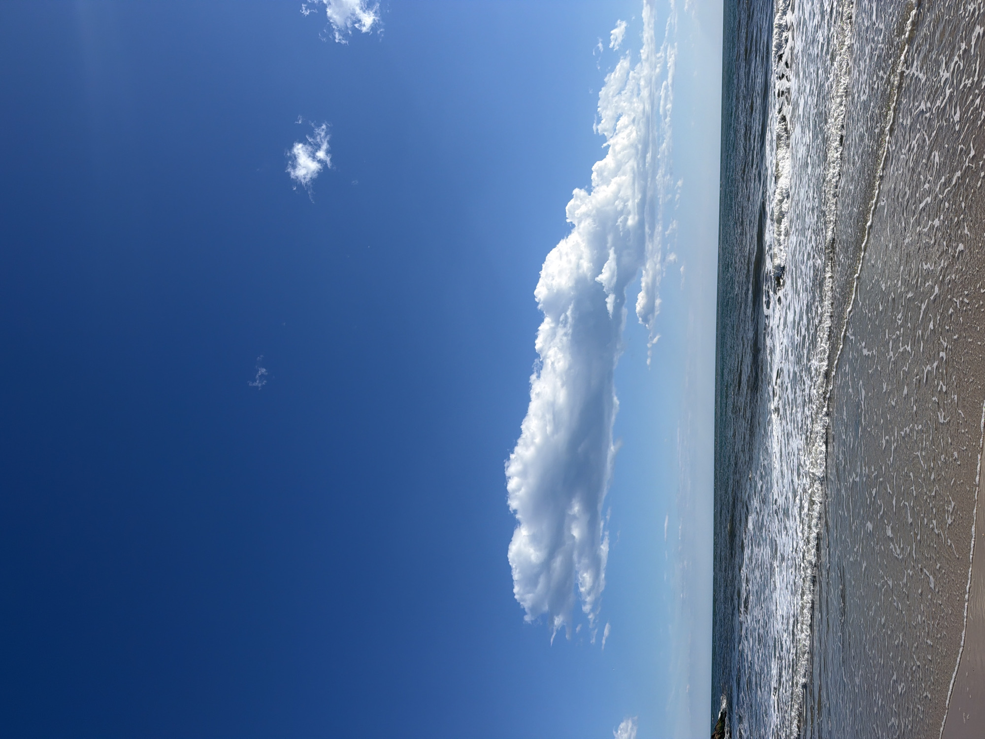 A bright, clear blue sky dominates the upper portion of the image, with a few scattered white clouds. The most prominent cloud formation is a long, horizontal bank of white, puffy clouds situated in the middle of the sky. Below the sky, the dark blue ocean stretches to the horizon. Small, white-capped waves are rolling towards the shore, with a foamy edge of breaking waves closer to the foreground. The very bottom of the image shows the wet sand of a beach, with receding water creating a shimmering, reflective surface. A dark, rocky structure juts out of the water on the far left side of the frame. The scene depicts a sunny day at a beach in Hampton Bays, United States of America. There are no people or animals visible in the image.