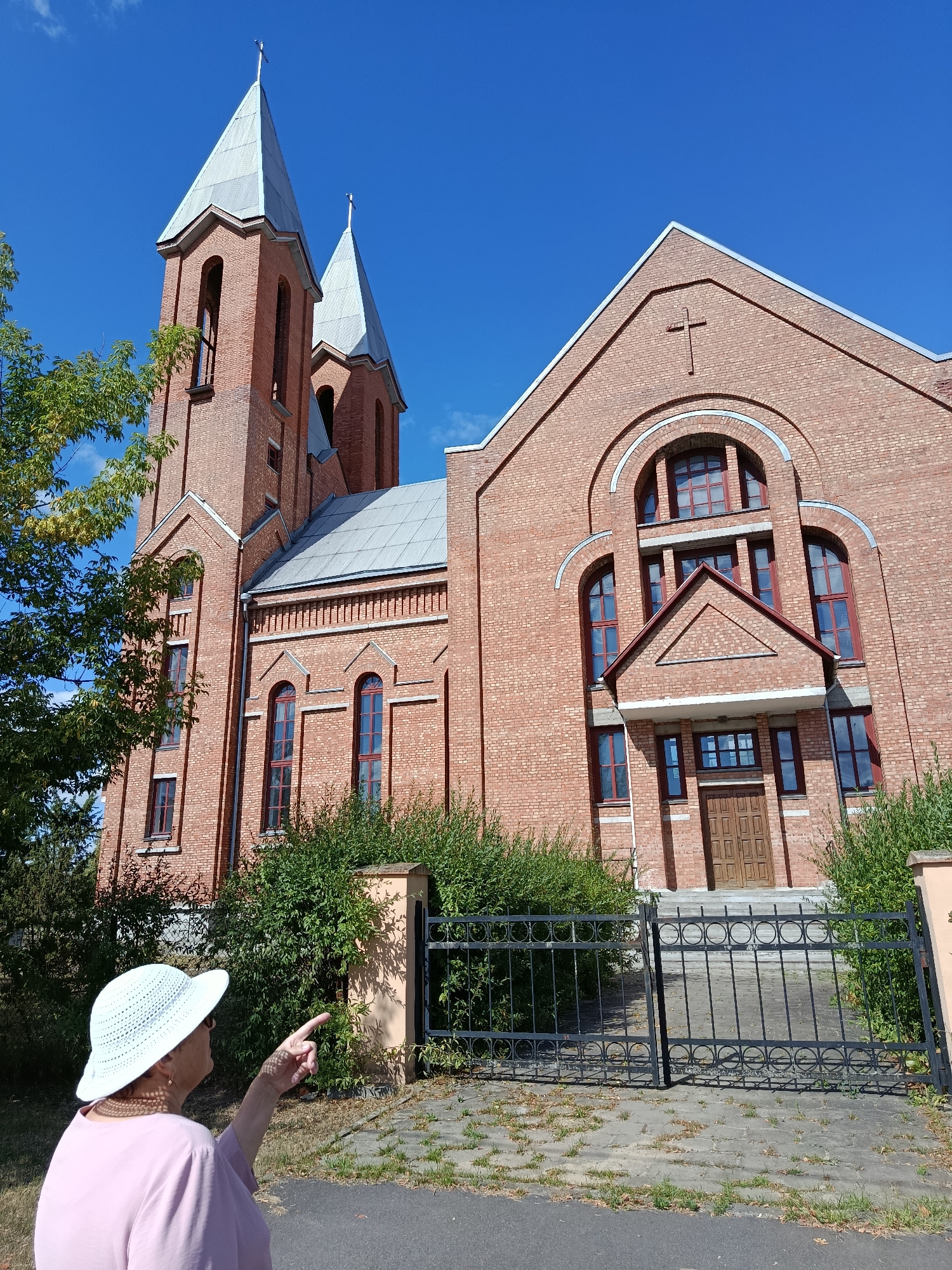 A medium shot photograph captures an elderly woman wearing a light-colored, wide-brimmed hat and a pink top, pointing towards a large brick church. The church features two tall spires, one taller than the other, both topped with crosses. The building is constructed from red bricks and has a large arched entrance with a wooden door. The scene appears to be set on a sunny day with a clear blue sky. The church is likely located in the city of Svietlahorsk, Belarus, given the architectural style often found in that region.