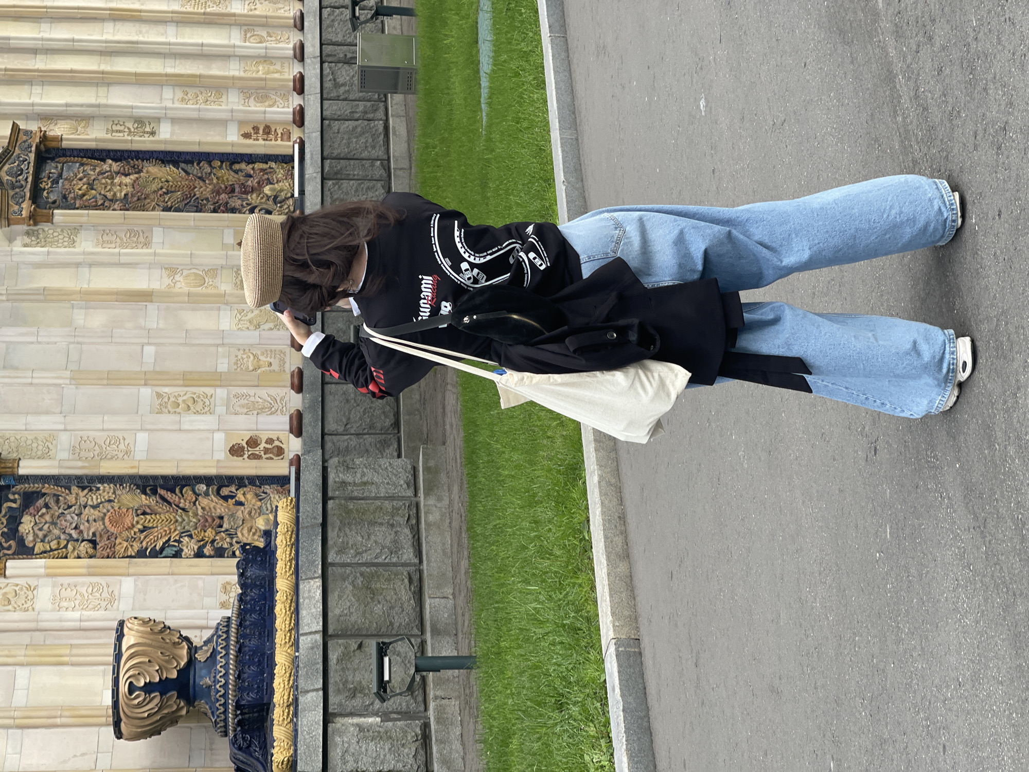 A person wearing a straw hat, a black and white long-sleeve shirt, light blue jeans, and white shoes is standing and taking a picture with a phone. The person also has a white tote bag over their shoulder and a black garment draped over their arm. The person is standing on a gray paved surface with a strip of green grass to their left. To the left of the grass is a stone structure with a tiled decorative panel. The location is Unknown, Unknown.