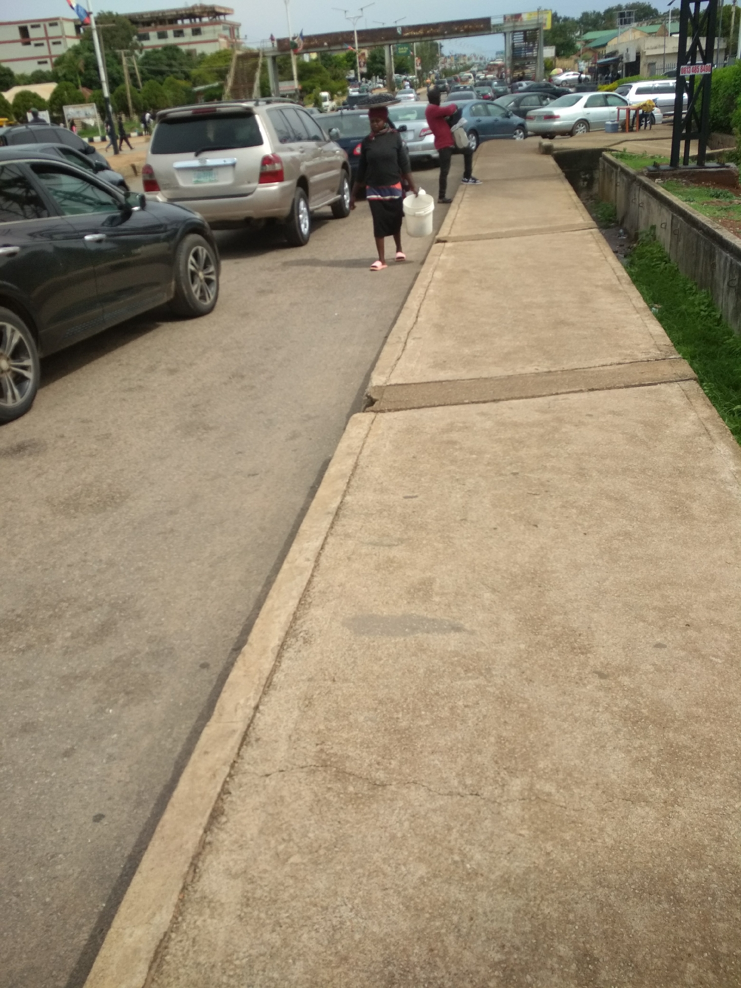 The image captures a daytime urban street scene in Jos, Nigeria, featuring vehicles, pedestrians, and infrastructure under an overcast sky.
In the foreground to the left, the front wheel and part of a black car are visible on the asphalt road. The road surface shows some dirt and minor wear. Parallel to the road, on the right, runs a concrete sidewalk composed of several slabs, with some visible cracks and a darker horizontal strip crossing it.
On the road in the middle ground, a light-colored (possibly beige or silver) SUV is prominently featured, seen from the rear, facing away from the camera. Other cars, including a blue sedan and several dark-colored vehicles, are visible further down the road, suggesting moderate traffic or parked cars.
Two main figures are present. One person, appearing to be a woman, walks on the edge of the road near the sidewalk, carrying a white bucket in her left hand. She is dressed in dark clothing and pink flip-flops. Further down the street, another person, possibly a man in a red shirt, stands near a vehicle or on the sidewalk. Other indistinct figures can be seen in the distance.
The background reveals multi-story buildings, lush green trees, and utility poles. An overhead gantry or bridge structure spans across the road in the distance. To the far right, a drainage ditch with concrete walls and some grass runs alongside the sidewalk. A black metal structure, possibly a pole or part of a sign, stands near the ditch.
Notable details include a phone number "0813 485 8490" visible on a billboard or sign in the far background to the right. The overall impression is one of a typical bustling street in a Nigerian city.