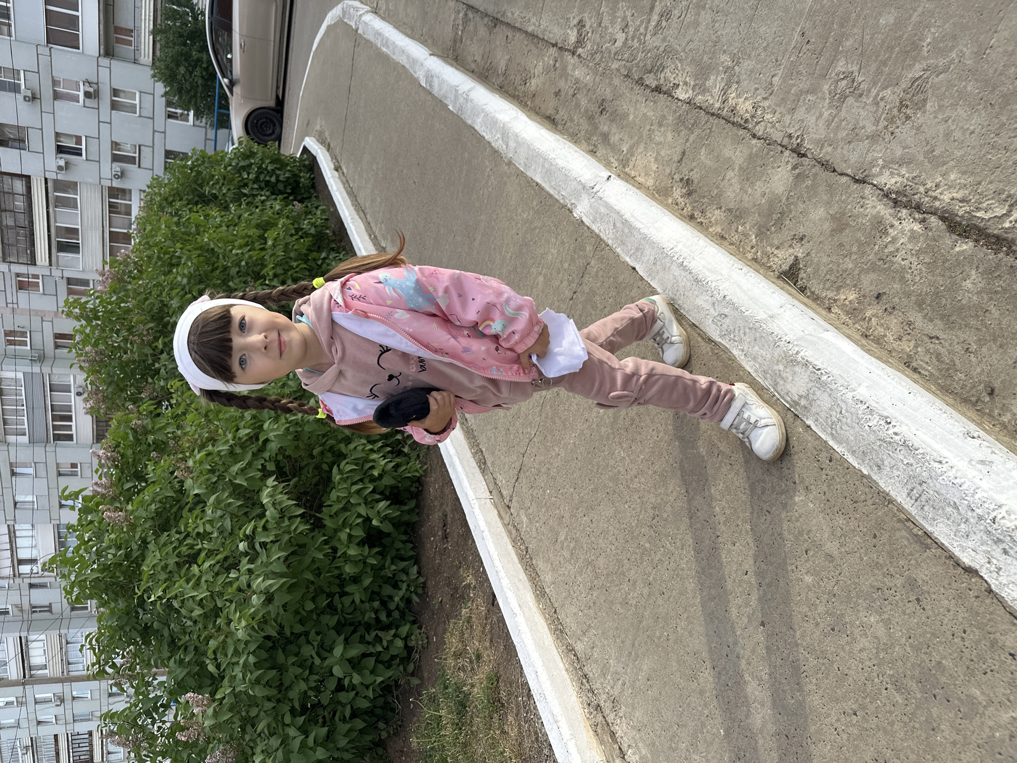 A young girl with pigtails stands on a concrete sidewalk next to a curb. She is wearing a pink tracksuit, white sneakers, and a white headband. She holds a black object in her hand. Behind her is a bush and a multistory apartment building. A car is partially visible in the background. The location appears to be in a residential area of Chaikovsky, Russia.