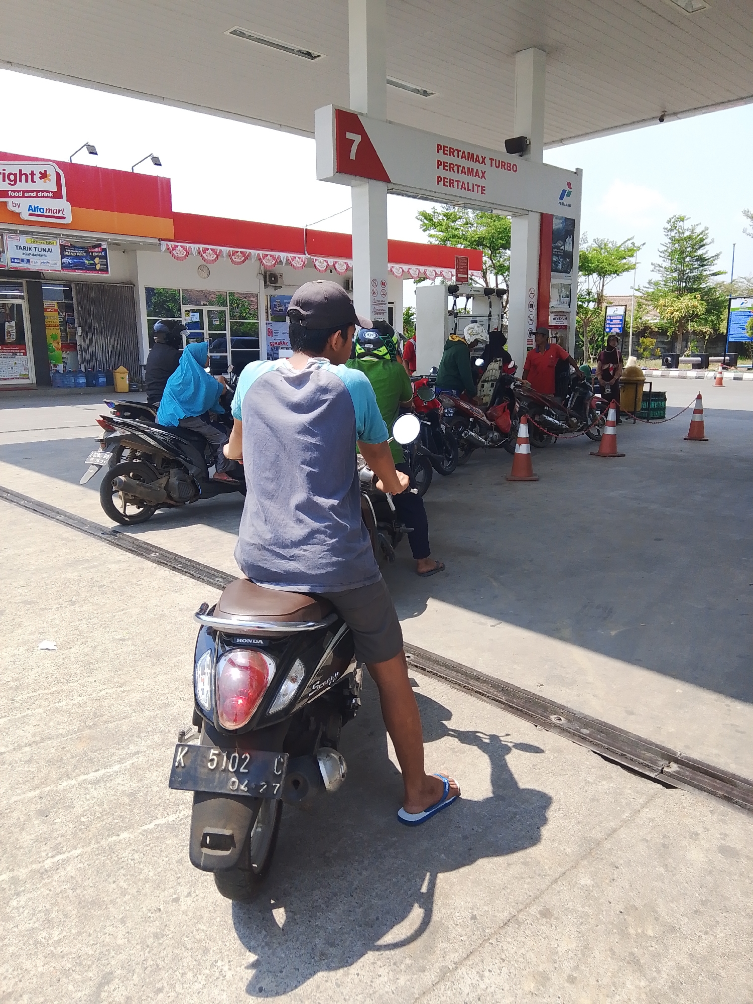 The image depicts a scene at a Pertamina gas station in Gedangan, Indonesia, on a sunny day. In the foreground, a person wearing a blue and gray t-shirt and dark shorts rides a black Honda Scoopy motorcycle. The motorcycle has a license plate reading "K 5102 C 04 27". Behind this motorcycle, another black motorcycle is visible with a rider wearing a blue headscarf and a dark jacket. Further back, several other motorcycles and individuals are queuing at the gas pumps. Red and white decorative banners hang under the station's canopy. The gas station appears to be attached to an Alfamart convenience store. The ground is paved concrete, and traffic cones are placed in various locations. Trees and other buildings are visible in the background. The overall atmosphere suggests a busy, ordinary day at a local gas station.