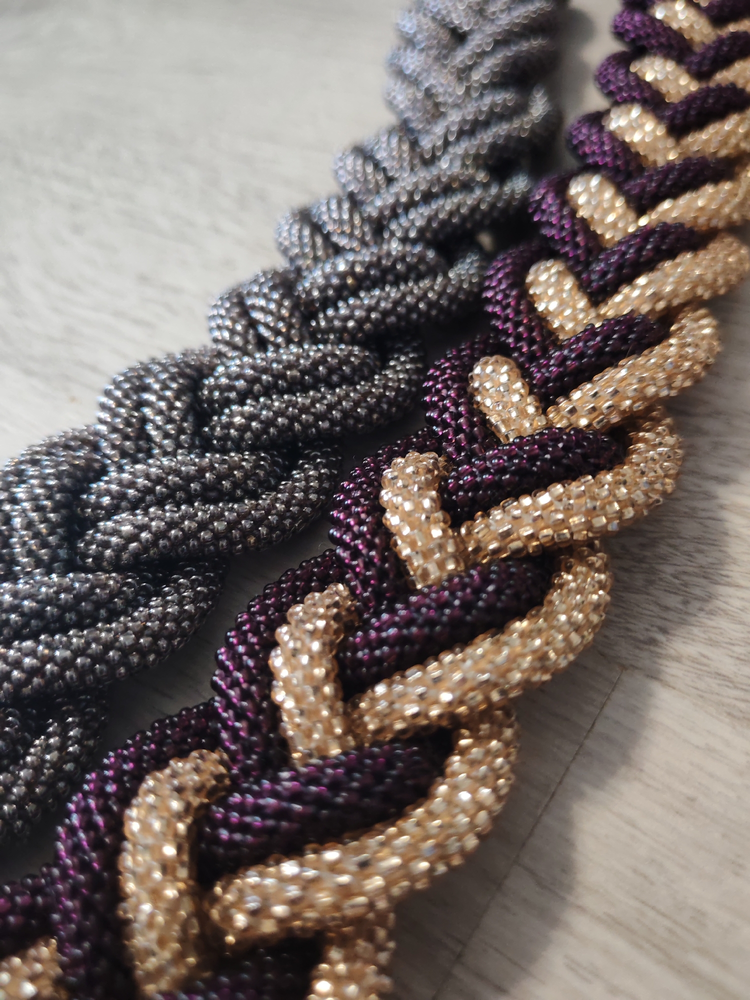 Two braided necklaces lie on a wooden surface. The necklace in the foreground is made of alternating purple and gold beads. The necklace in the background is made of silver beads. The lighting is artificial, casting shadows on the wooden surface. The scene appears to be indoors, possibly a home or studio. There are no people present in the image. The photograph is taken from a high angle, looking down at the necklaces. The focus is on the texture and intricate detail of the beadwork.