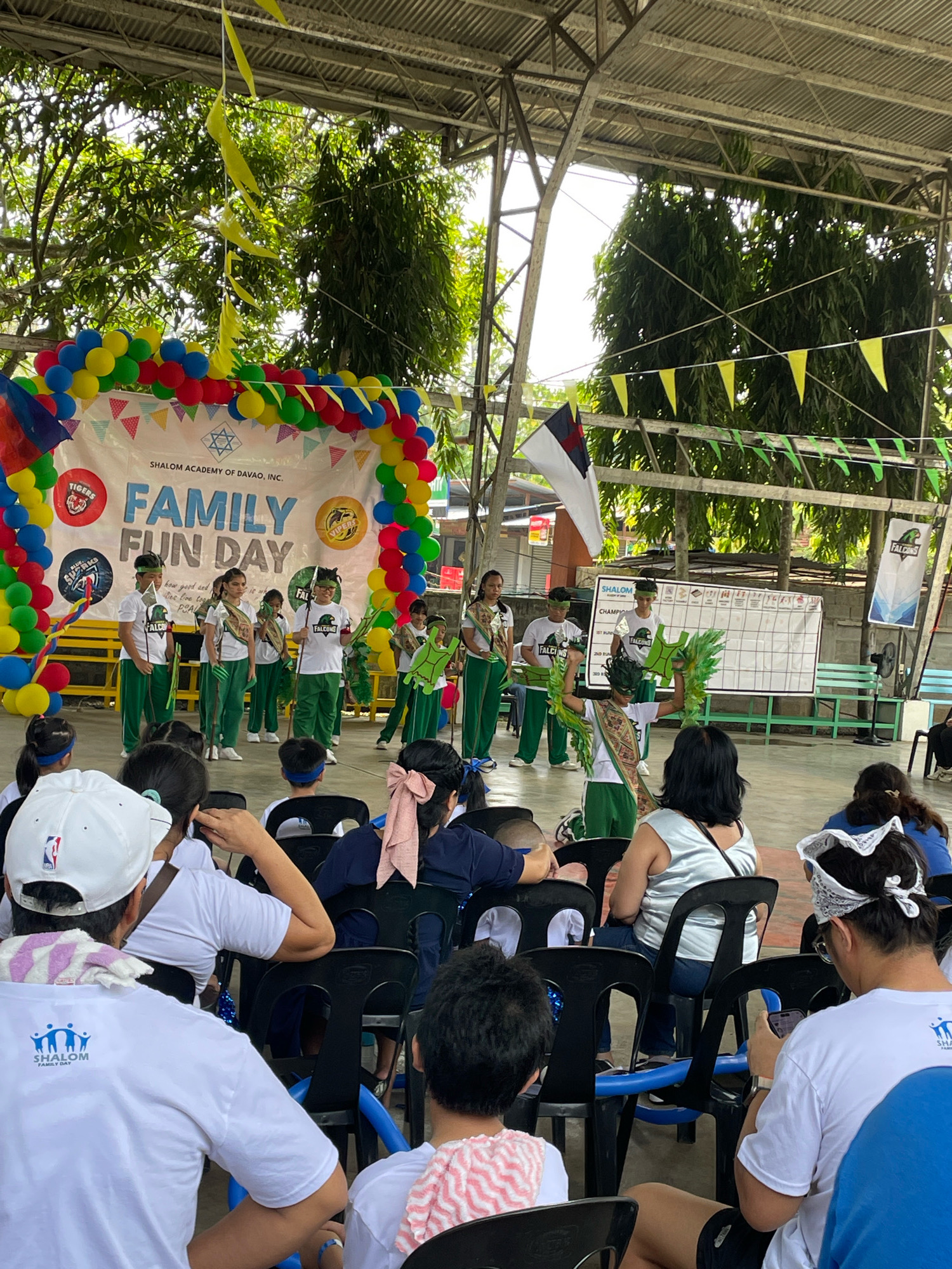 This image captures an outdoor event at Shalom Academy of Davao, Inc., located in Davao City, Philippines. The scene appears to be a "Family Fun Day," as indicated by the prominent banner. In the background, a stage is set up under a covered structure, adorned with a colorful balloon arch and pennant banners. Several children dressed in green and white uniforms are on stage, some holding props like green leafy accessories and what appears to be a turtle costume. A group of attendees, mostly children and adults, are seated on plastic chairs in the foreground, facing the stage. The environment is characterized by lush green trees visible beyond the covered area, suggesting an open-air or semi-outdoor venue. The overall atmosphere seems festive and community-oriented.