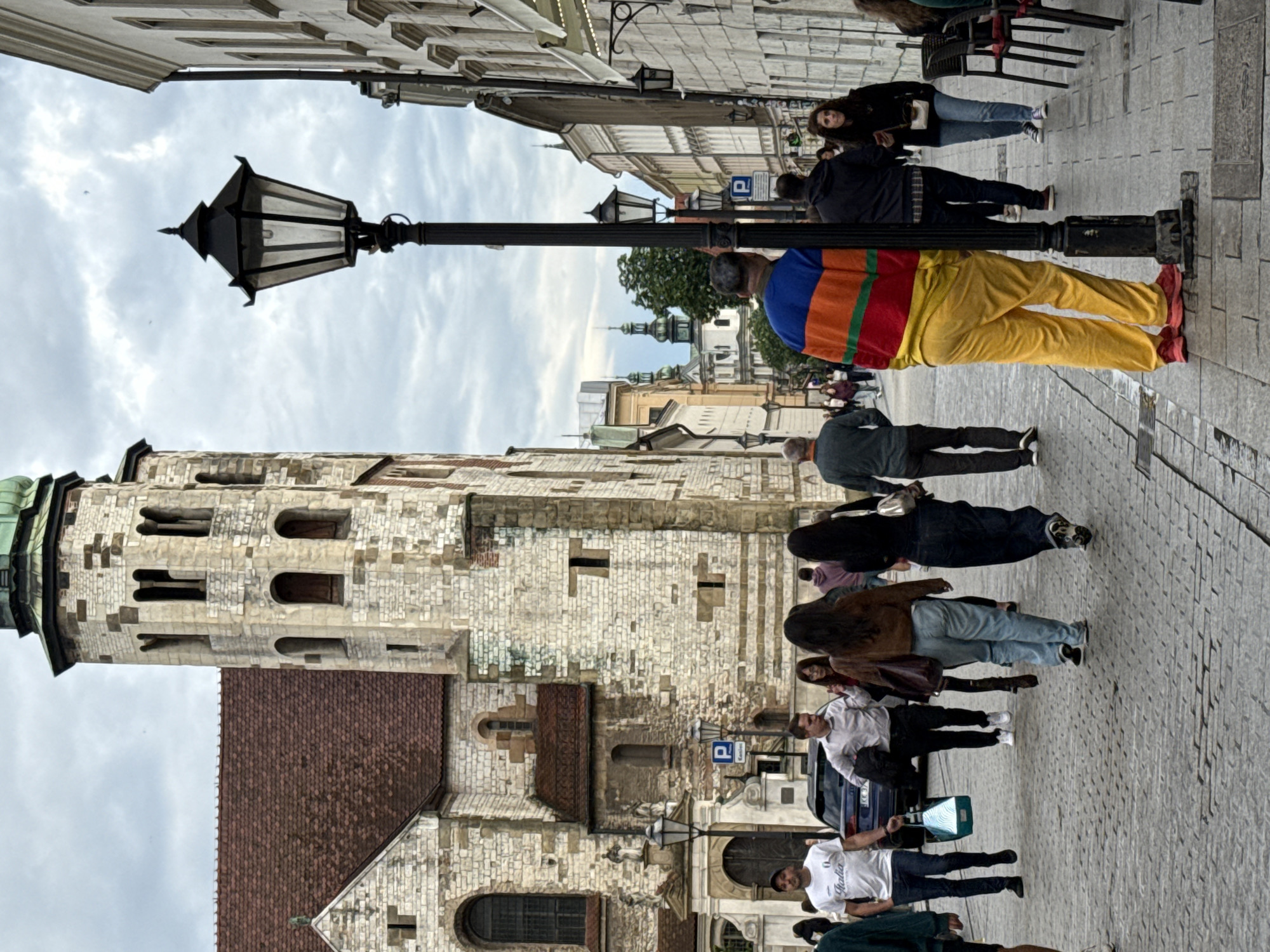 The image depicts a street scene in Krakow, Poland, likely in the Old Town. In the foreground, a person wearing a brightly colored striped sweater and yellow pants stands near a lamppost. To the left, a stone building with a red tiled roof and a tall tower dominates the background. Several people are walking on the cobblestone street in front of this building. To the right, a row of buildings with cream-colored facades lines the street, some with awnings and outdoor seating. The sky is overcast. The scene captures a moment of daily life in a historic European city.