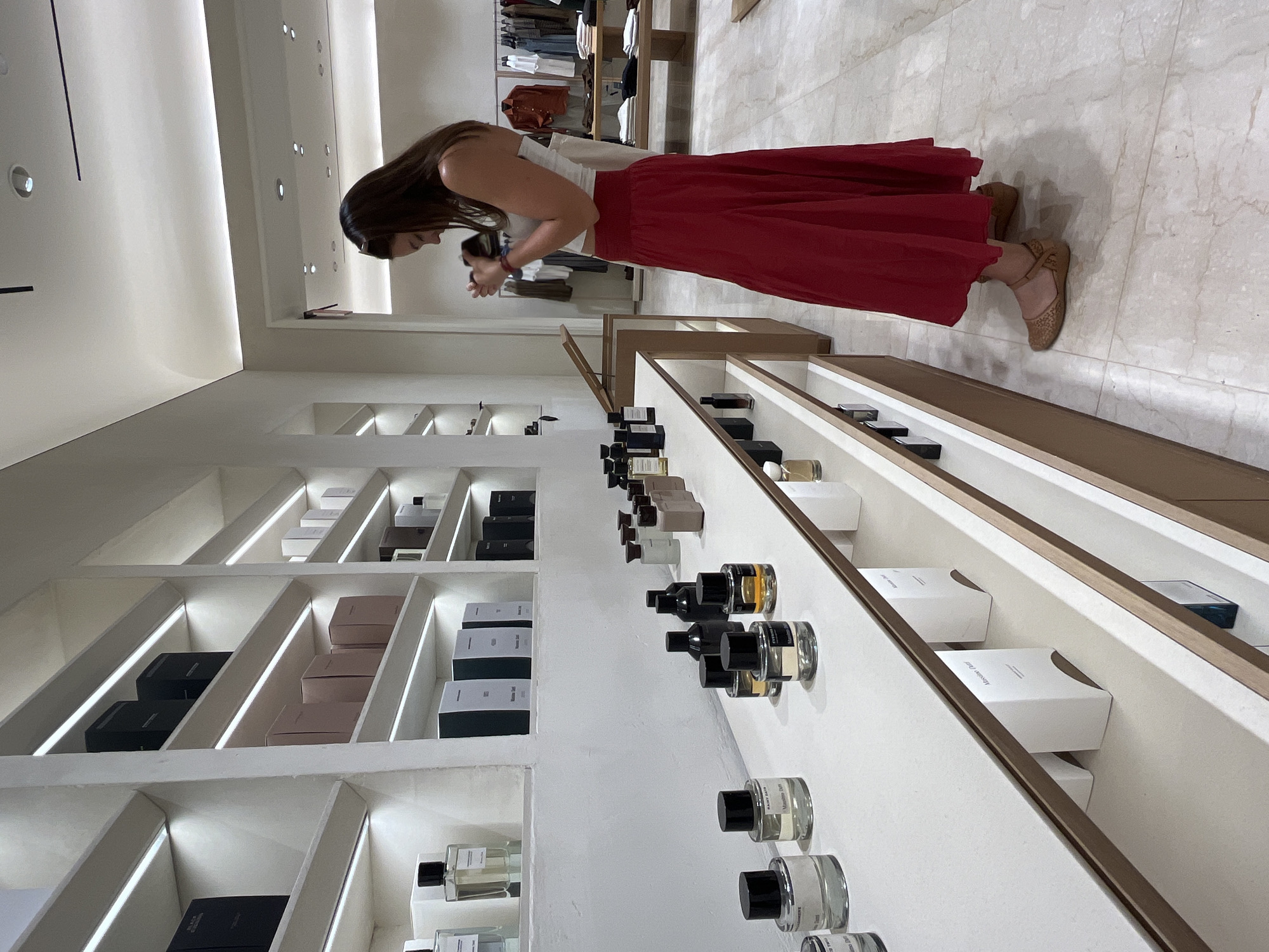 A woman with long brown hair is looking at a shelf filled with perfume bottles and boxed perfumes in a well-lit retail store. The shelves are white with recessed lighting. The woman is wearing a white top and a long red skirt. She is wearing brown flat shoes. The floor is tiled. The store appears to be a high-end establishment, as there are clothes displayed in the background. There is no evidence to identify Puebla, Mexico as the location.