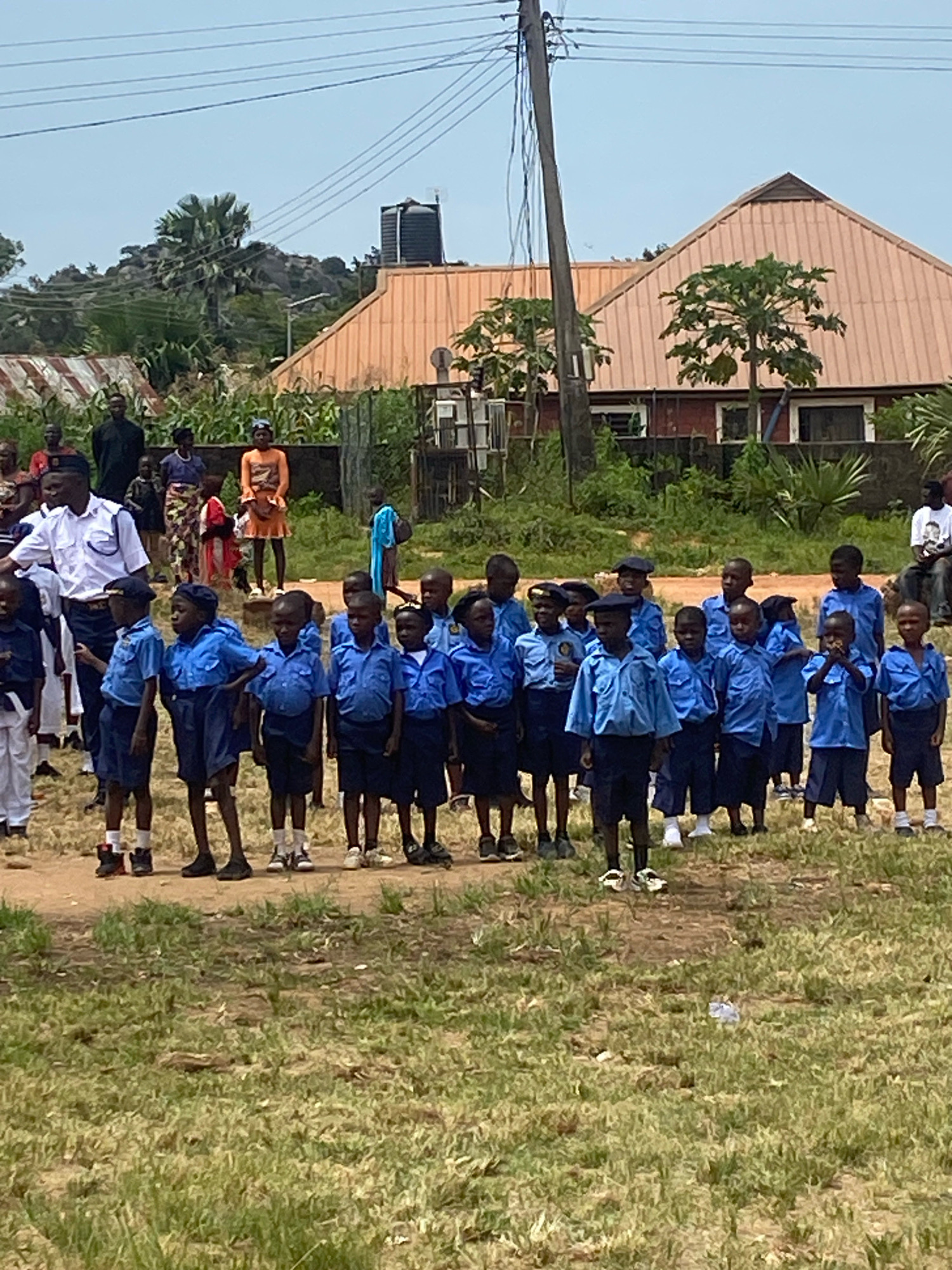 In Binchi, Nigeria, a group of young students dressed in blue uniforms stand in formation outdoors. Some students wear blue shirts and shorts, while others wear blue shirts and long pants. Several students have blue caps. A man in a white uniform, possibly a teacher or school staff member, stands to the left of the students. Behind the children, adults and other children are visible in the background, some wearing traditional Nigerian clothing. The environment is characterized by dry, dusty ground with patches of dry grass. In the background, buildings with corrugated metal roofs and trees are present, under a clear sky with visible power lines. The scene appears to be a school event or assembly.