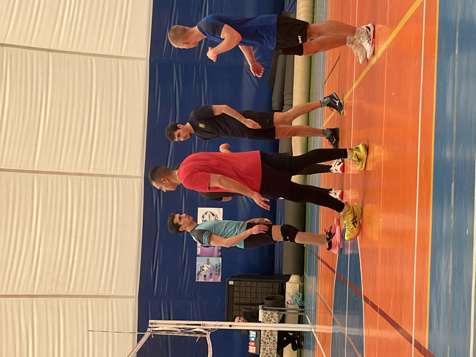 The image shows four volleyball players standing on a volleyball court, presumably indoors. They are lined up and appear to be facing the same direction. The court has orange and blue sections separated by yellow lines. A volleyball net is partially visible in the foreground. Behind the players is a blue wall with several posters. The background also includes a white, tent-like structure, likely the ceiling of the indoor facility. A scoreboard is visible. The exact location in Sofia, Bulgaria cannot be determined from the image.