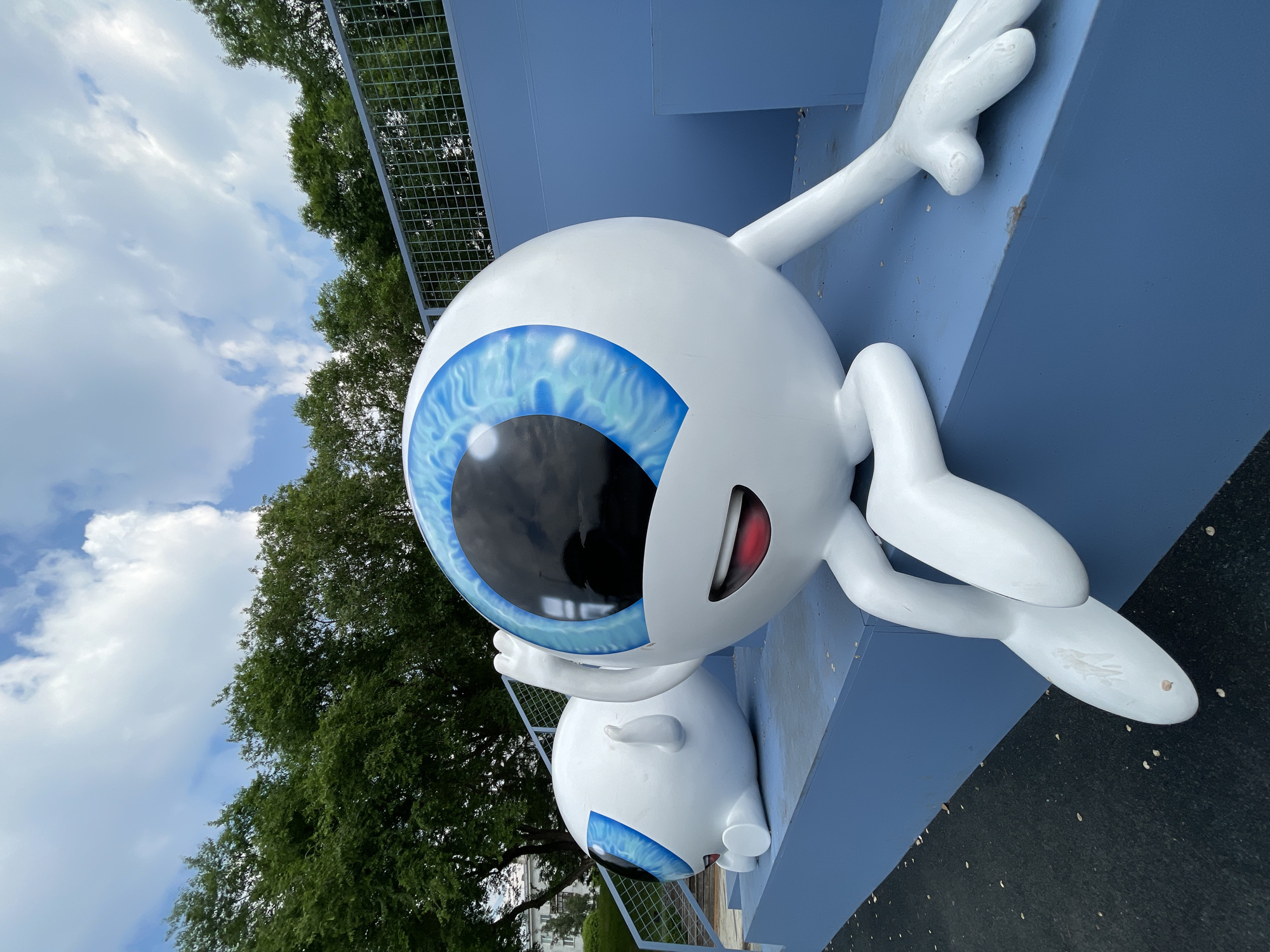 Two large, white, cartoon-style eyeball sculptures are situated outdoors on a bright blue tiered platform. The sculpture in the foreground is larger and positioned to the right, facing left. It has a bright blue iris with black pupils, a smiling red mouth, and white arms and legs. One arm is raised in a wave-like gesture, and its legs are crossed. The sculpture in the background is smaller and lies on its side to the left, also featuring a blue iris and black pupil, with its arm resting beside its head. The platform is surrounded by green trees and a metal fence, with a cloudy sky overhead. The sculptures are located in the Park of the Eye in Krasnoyarsk, Russia.
