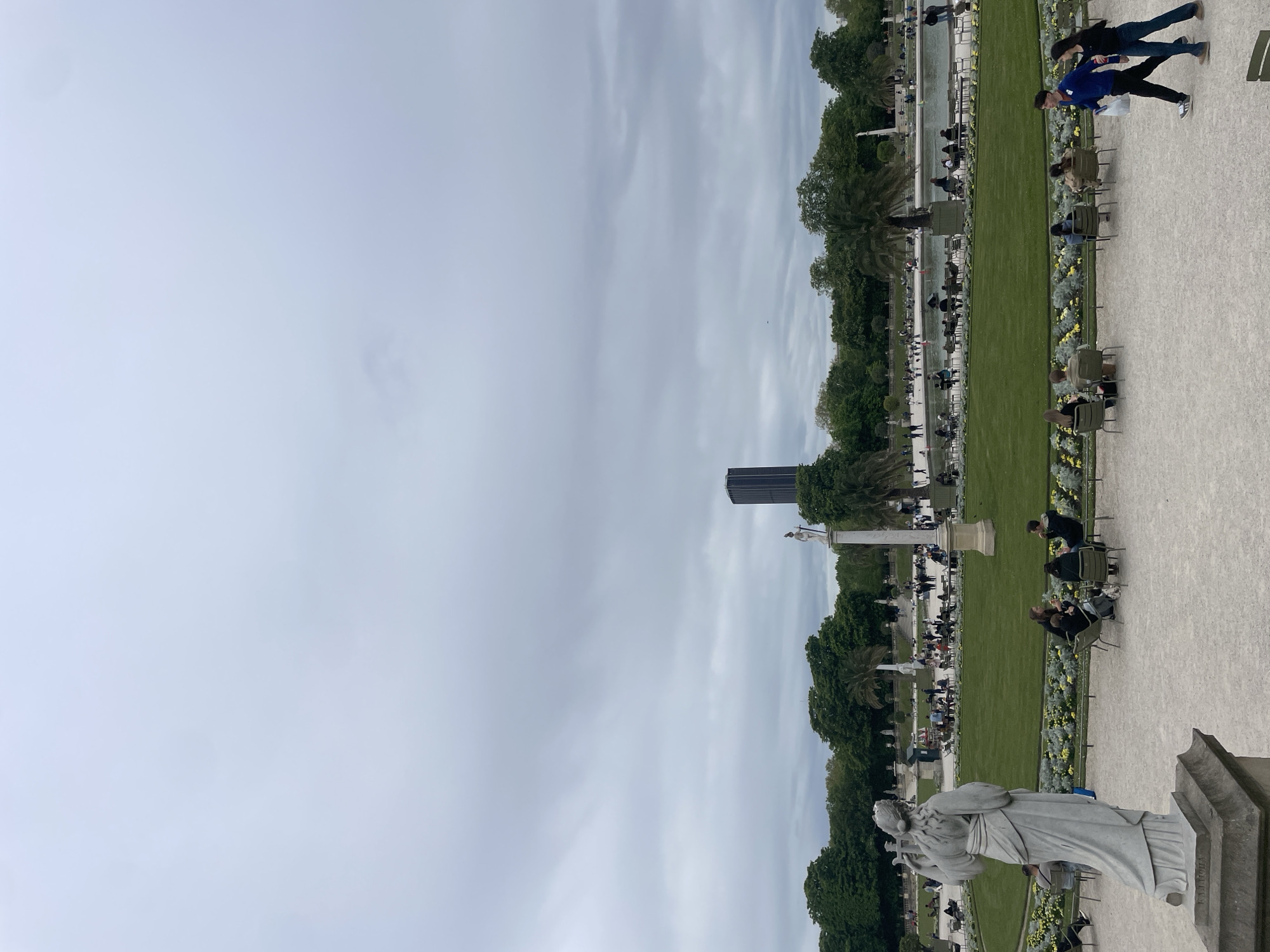 The photograph depicts a scenic view of the Tuileries Garden in Paris, France, characterized by a blend of natural and architectural elements. A classical statue stands prominently in the foreground, crafted from stone, depicting a draped figure. Extending behind the statue is a meticulously maintained green lawn, divided by gravel pathways. Along the pathways, individuals sit in rows of dark green chairs, spaced intermittently with planted yellow flowers. In the background, a modern high-rise building is visible amidst the trees. Overhead, the sky is overcast with gray clouds. People are seated, walking and interacting in the park.