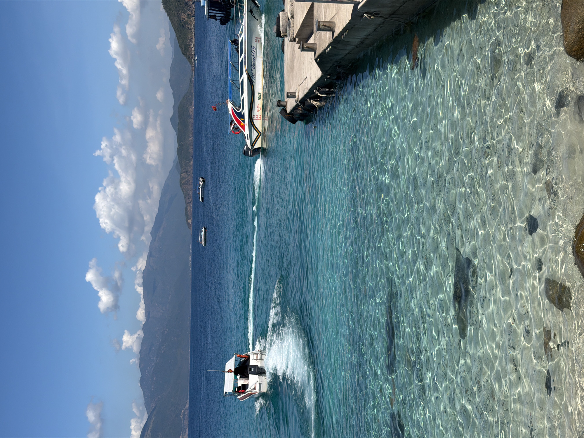 The scene depicts a sunny day in Nha Trang Ward, Vietnam. The clear turquoise water is close to a concrete pier. A large white boat is docked at the pier. Three smaller boats are visible in the sea. One is moving at speed, creating a wake. Mountains are in the background. The sky is blue with puffy white clouds.