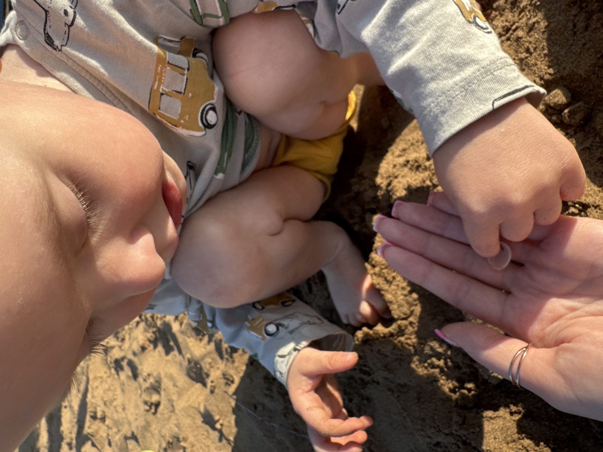 The image shows a close-up, overhead view of a baby and an adult's hands on a sandy surface. The baby is wearing a light gray onesie with a pattern of yellow and green toy trucks. The baby's face is partially visible on the left, with soft skin and wispy eyelashes. The baby's legs and torso are visible, with the baby wearing yellow shorts underneath the onesie. One of the baby's hands is clenched, while the other is open and reaching towards the adult's hand.
The adult's hand is in the lower right portion of the frame, with painted pink fingernails and a thin, gold ring on the thumb. The adult appears to be holding a small, round object, possibly a shell or a stone, between their thumb and index finger. The background is composed of coarse sand, suggesting an outdoor setting like a beach or sandbox. The lighting indicates it is daytime, with shadows cast on the sand. The overall scene evokes a sense of gentle interaction and play between the baby and the adult. There is no visible text in the image.