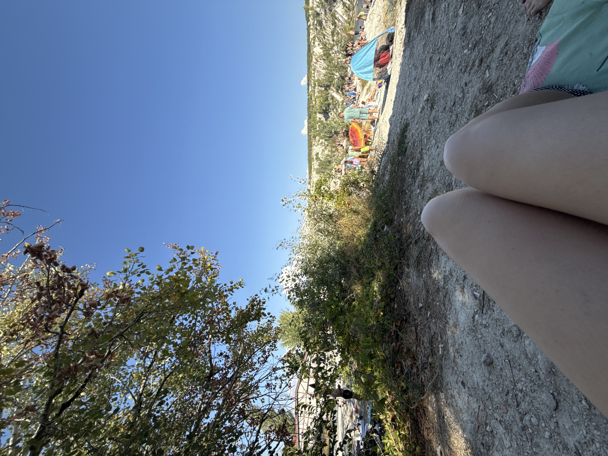 A wide-angle, ground-level shot captures a sun-drenched day at a beach or quarry in Скалистовское сельское поселение, Ukraine. The foreground is dominated by the legs and thighs of an unseen person, positioned as if relaxing on the ground. The ground itself is a mixture of sand, small stones, and dry foliage.
Beyond the foreground, the scene opens up to a populated area. Lush green trees and bushes line the left and central portions of the frame, with a building visible through a gap in the foliage. Further back, a white, chalky embankment rises, characteristic of a quarry, with more greenery and trees dotting its slopes.
The beach area is bustling with activity. Numerous people are scattered across the sand, some sunbathing, others standing and interacting. Several beach umbrellas and tents are set up, including a prominent blue tent on the right side of the frame. An orange inflatable object, possibly a float or raft, is also visible among the beachgoers. The sky is a clear, vibrant blue, indicating fair weather. The overall impression is one of a lively summer day at a recreational spot.