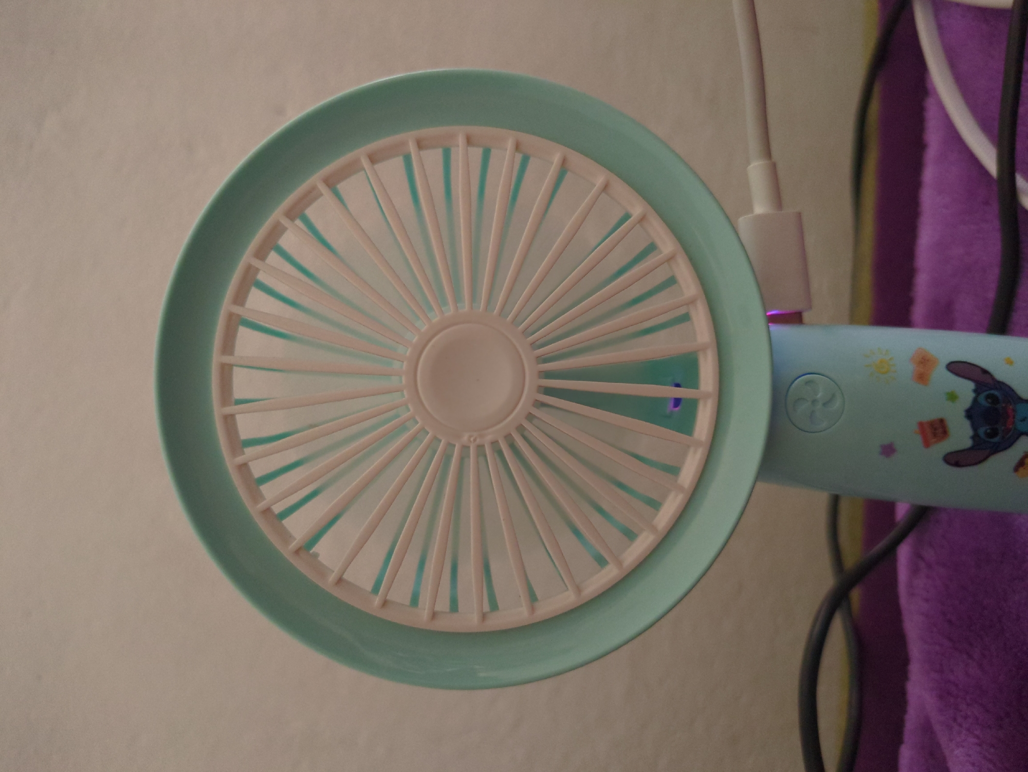 A teal and white portable fan is displayed prominently in the foreground against a neutral, textured background. The fan features a circular grille with numerous white spokes radiating from a central hub. A light purple glow emanates from beneath the fan blades. The base of the fan is also teal and adorned with colorful cartoon decals, including a character resembling Stitch from Disney's Lilo & Stitch. A white USB charging cable is plugged into the side of the fan's base. To the left of the fan, a dark grey cable is visible, resting on a soft, purple fabric surface. The scene is static and does not depict any action or interaction. The location is identified as Rusiec, Poland.