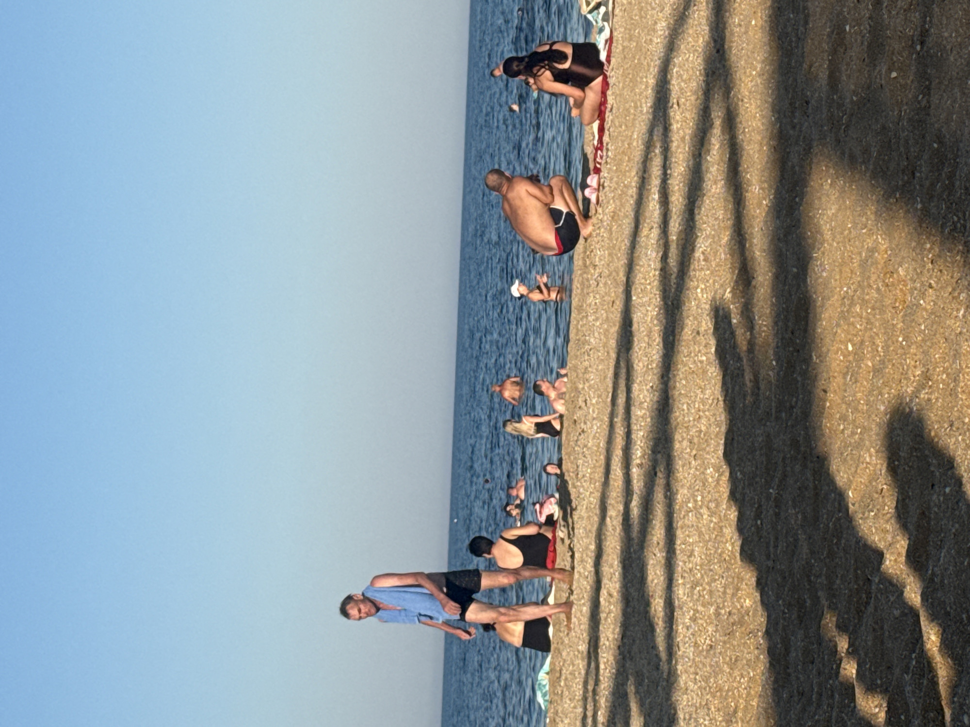 The image depicts a sunny day at a beach in Ukraine. The foreground is a sandy beach with shadows cast by what appears to be a palm tree. The middle ground shows several people in the shallow water of the sea, some are swimming, while others are standing or sitting. In the background, the sea stretches to the horizon under a clear blue sky. On the left side of the image, a man in black swim shorts and a blue towel draped over his shoulder stands on the sand, facing left. To his right, a woman in a black swimsuit sits on the sand. Further to the right, a man in black swim shorts with red trim is crouched, facing a woman in a black swimsuit who is sitting. Several other people are visible in the water. The overall scene is one of a relaxed day at the beach.