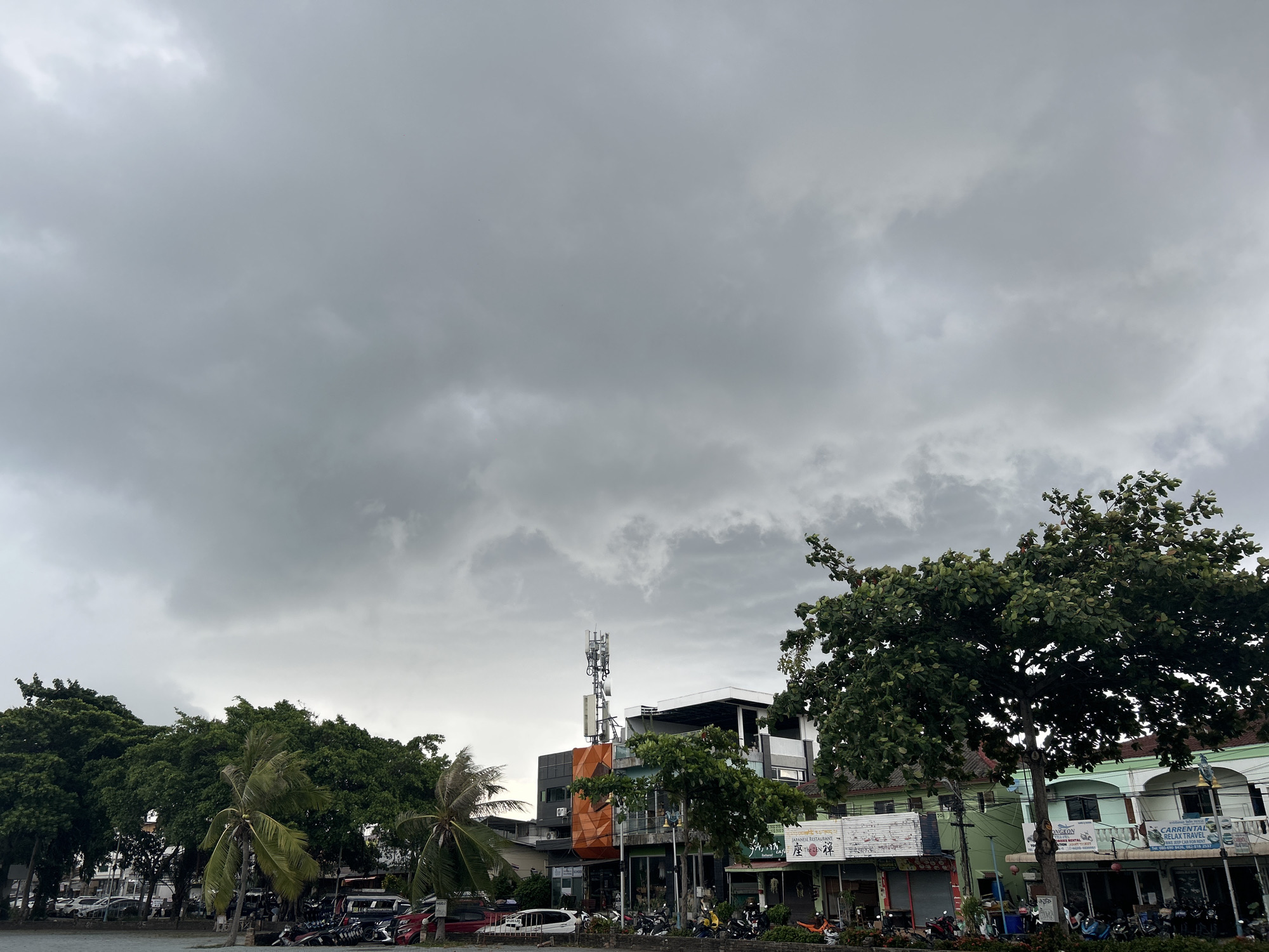 The image depicts an overcast day in a tropical or subtropical town, consistent with ตำบลเกาะพะงัน, Thailand. The sky is largely covered by heavy, grey clouds, suggesting gloomy weather, possibly preceding rainfall.
In the foreground and midground, a street or waterfront area is lined with a variety of multi-story buildings and abundant green vegetation. Lush trees with dense foliage, including several palm trees, are prominent on both sides of the frame. Parked cars, numerous scooters, and motorbikes are visible along the street.
A central building exhibits a more modern architectural style, featuring an orange-paneled facade on its lower level and a grey upper structure. A large cell tower with multiple antennas stands conspicuously on its rooftop. To the right, other buildings display varying colors and architectural elements, including balconies and shopfronts.
Visible text on signs provides specific location cues. A sign on one building clearly advertises a "JAPANESE RESTAURANT" with Japanese characters (座禅) and possibly "ZAZEN CAFE". Further right, another sign reads "C.ARRENTAL RELAX TRAVEL". A partially visible sign on a tree also shows "ONGRON".
No people are clearly visible in the scene, which appears static. The heavy cloud cover sets a subdued mood, typical of an overcast day. The combination of tropical plant life, diverse buildings, and specific signage firmly places the setting in a town in Southeast Asia, identified as Koh Phangan, Thailand.