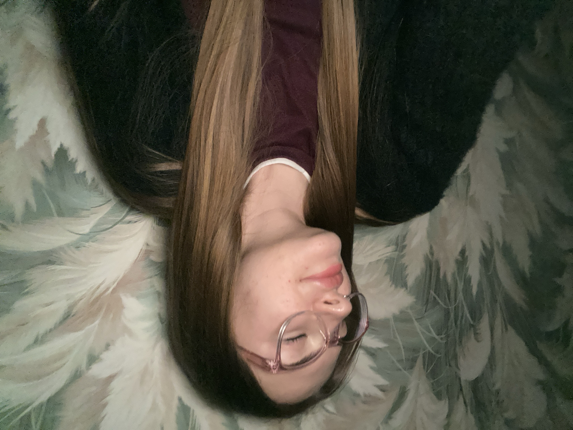A person is lying down in what appears to be an indoor setting. The individual has long, light brown hair fanned out around their head. They are wearing a dark maroon long-sleeved top with a white trim along the collar, and are partially covered by a dark, possibly black, fuzzy garment or blanket. The person wears glasses with thin, light-colored frames, and their eyes are closed or mostly closed, suggesting a relaxed state. Their lips are a soft pink, and a few freckles are visible on their nose and cheeks.
The surface they are lying on features a distinctive pattern of light, feathery or stylized foliage designs in shades of white, light green, and teal. This pattern is spread across the background, suggesting it could be a bedspread, blanket, or decorative fabric. The lighting is soft and diffused, creating a calm atmosphere. No specific location cues are visible beyond being an indoor space. There is no visible text in the image. The overall impression is one of peace and relaxation.