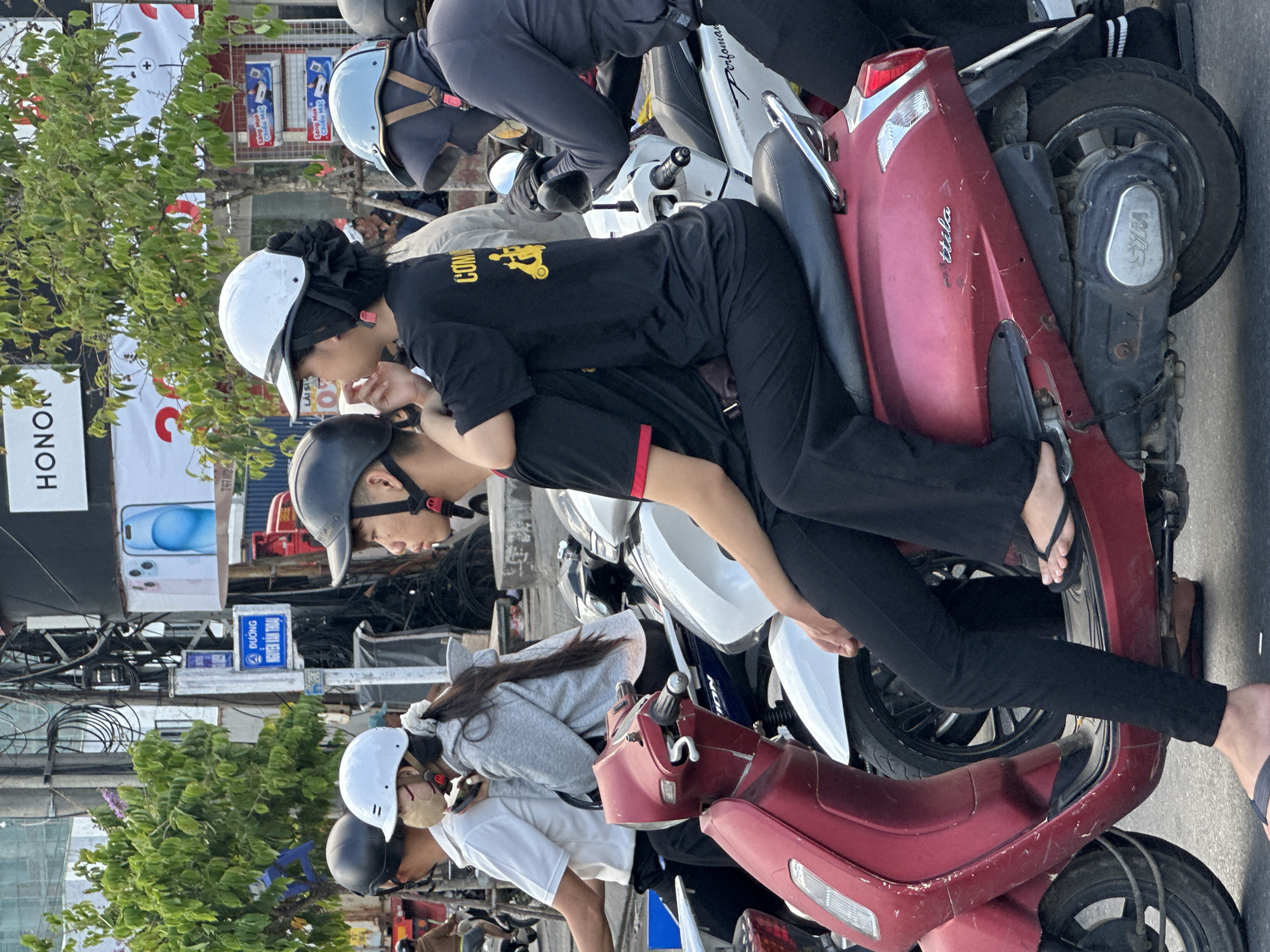 This image captures a street scene in Da Nang, Vietnam, likely during the daytime. The primary subjects are several individuals on and around motorbikes, a common mode of transportation in the region.
In the foreground, two people are seated on a red motorbike. One person, wearing a black t-shirt with yellow lettering that reads "COM" and black pants, is positioned behind the other, who is wearing a white helmet. Their posture suggests they might be a passenger embracing the driver or simply sharing the seat. The driver is wearing a black helmet.
Behind them, another red motorbike is visible, partially obscured. Further back, a white motorbike with a rider wearing a blue and white helmet is also present. The background features a mix of elements typical of a Vietnamese street: electrical wires strung across poles, buildings, signage, and lush green trees.
One sign prominently displays the word "HONOR," likely an advertisement for a technology brand. Other signs show Vietnamese text, including "ĐƯƠNG" and "NGUYỄN VĂN THOẠI," indicating street names, and what appear to be promotional messages like "Sông Mua Chiều Má."
The overall atmosphere is bustling, reflecting daily life in a busy urban environment. The presence of helmets suggests adherence to traffic regulations or a general safety awareness among the motorbike riders. The lighting and shadows indicate it's likely daytime, possibly late afternoon given the angle of the sun. The scene is a snapshot of everyday transit and urban activity in Da Nang.