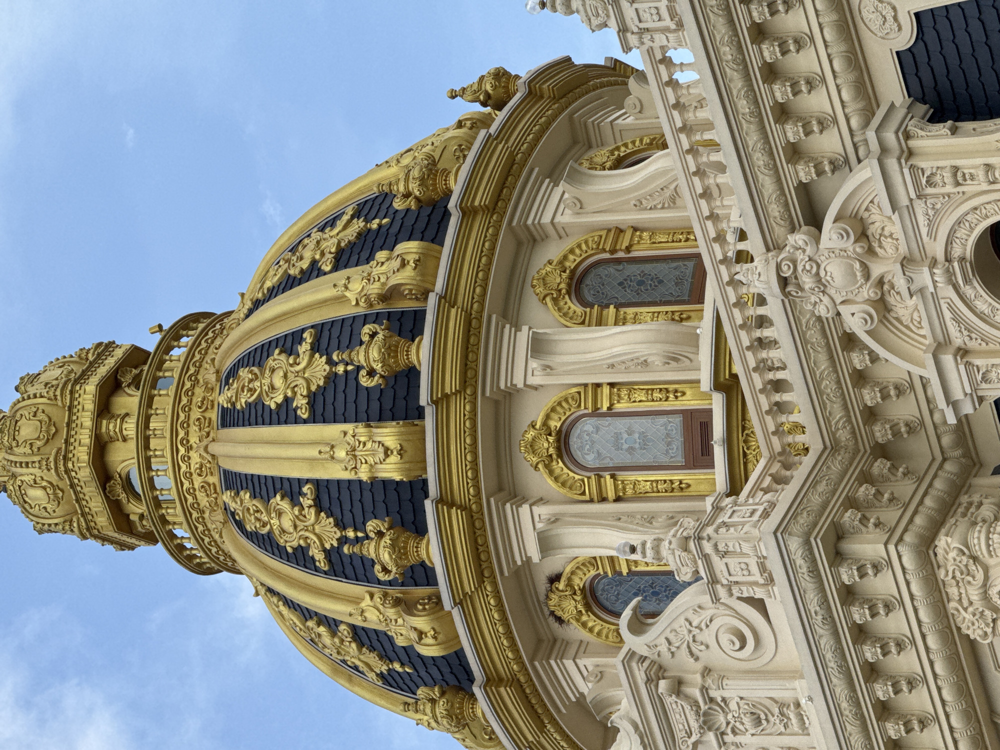 The image shows a close-up of a golden dome and the upper facade of a building, likely a religious or historical structure. The dome is adorned with intricate gold detailing against a dark, tiled surface, and is topped with a golden spire. Below the dome, the building's exterior features elaborate cream-colored ornamentation, including decorative moldings, arched windows with stained glass, and a balustrade. The sky, with a few wispy clouds, is visible in the background. This architectural style is characteristic of the Dinh Ba historical site in Da Lat, Vietnam, known for its French colonial influences and ornate design. There are no people or animals visible in the image.
