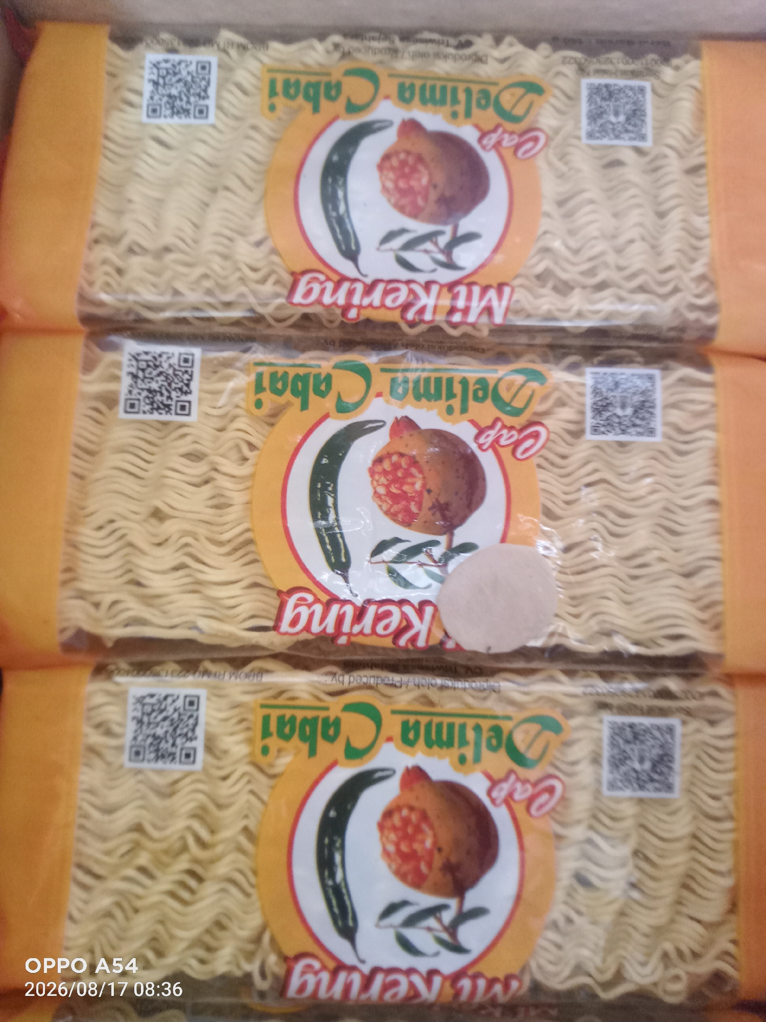 The image shows three packages of instant noodles stacked vertically. Each package has a yellow band on the side. The front of each package features a circular logo with a picture of a pomegranate and a chili pepper. Text on the logo reads "Cap Delima Cabai Mi Kering". The noodles inside are visible through the clear plastic packaging. The background is out of focus, but it appears to be a store or market shelf. The bottom left corner of the image has a timestamp and model name overlay: "OPPO A54 2026/08/17 08:36". There are no people or animals in the image. The location is not specified in the image, and therefore cannot be determined to be Jambi, Indonesia.
