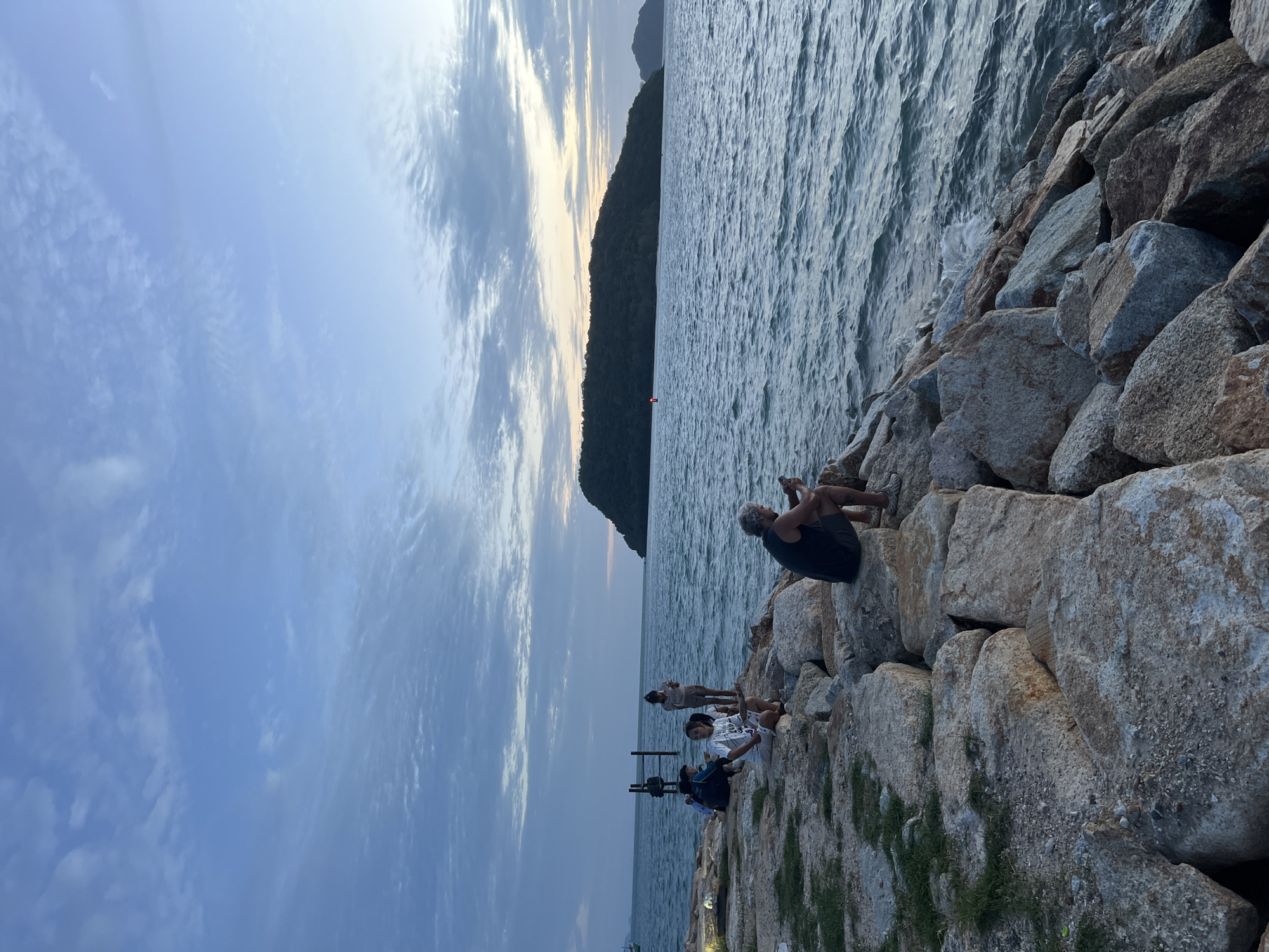 A group of people sit on large rocks along the edge of a body of water. The water is choppy and a dark blue, with small waves lapping against the rocks. In the distance, a lush green island is visible, its silhouette outlined against a soft, cloudy sky. The sky itself is a pale blue, with streaks of white clouds that suggest the transition from day to evening.
Among the people, a man with gray hair sits on a rock, looking down at his phone. To his left, a woman in a white t-shirt and shorts sits on the rocks, also looking down, possibly at her phone. Two other individuals are seated further back, their postures relaxed.
The rocky shoreline is covered in large, rough-hewn stones of various sizes, with patches of sparse green grass growing between them. A dark, possibly wooden, structure stands in the water to the left of the frame, with a few horizontal beams. A red buoy is visible in the water near the island. The overall impression is a serene coastal scene, with people enjoying the view and the quiet atmosphere.