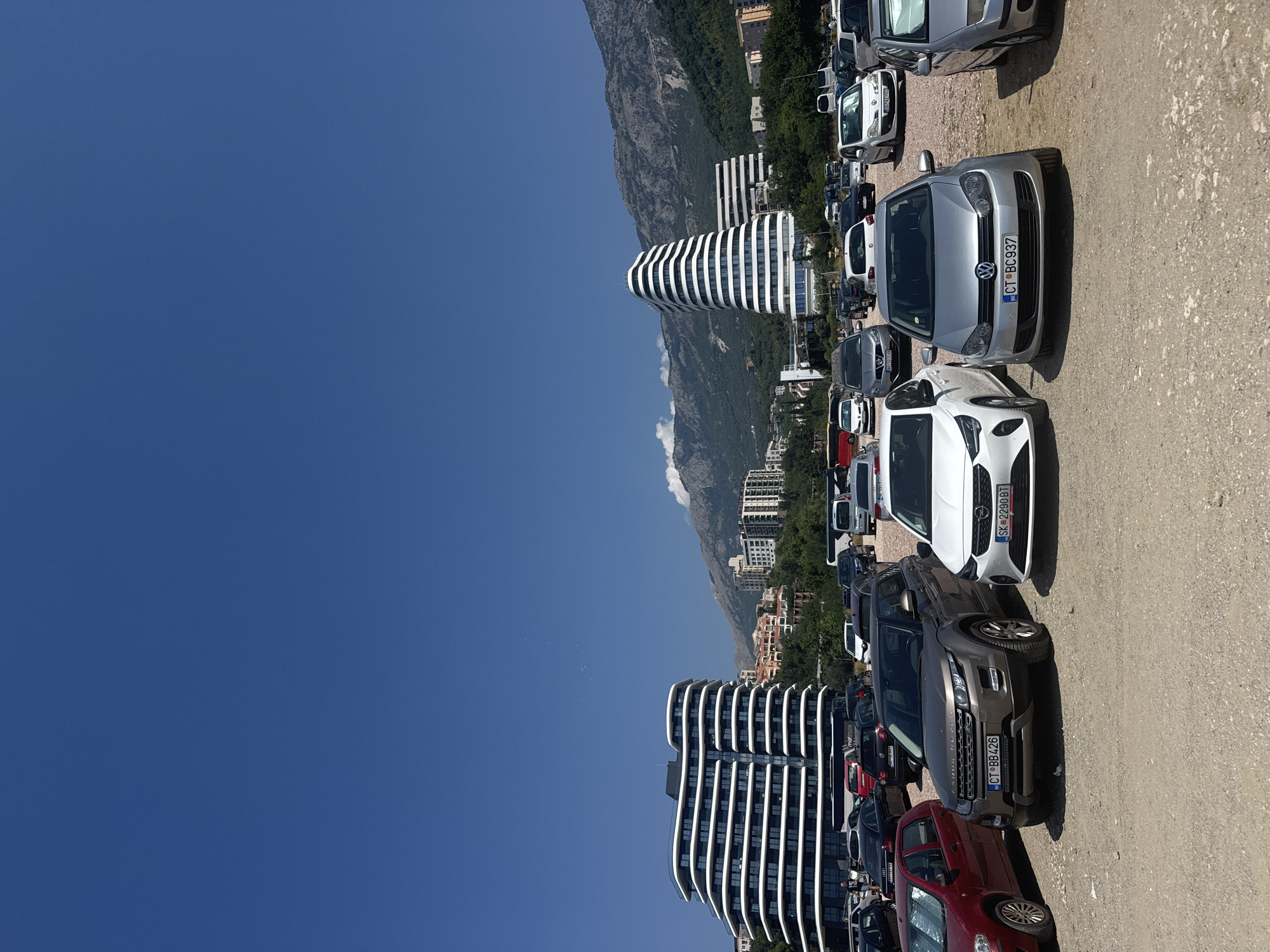 A wide shot captures a parking lot filled with cars under a clear, bright blue sky. In the background, modern buildings with curved facades stand against a backdrop of lush green trees and imposing, rocky mountains. The parking lot itself is unpaved, composed of gravel and dirt, with various vehicles parked in rows. The foreground is dominated by several cars, including a dark grey Range Rover, a white Opel Corsa, and a silver Volkswagen Golf. The overall scene suggests a sunny day in a coastal or mountainous urban area.