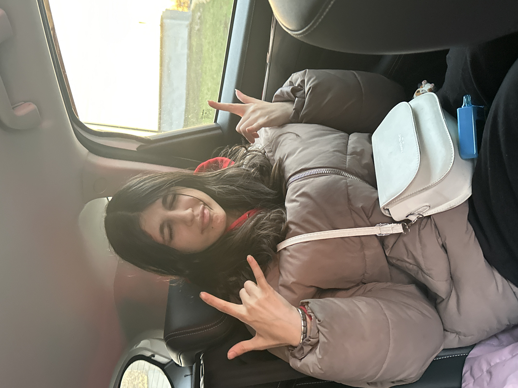 A young woman with long dark hair is seated in the back of a car, wearing a puffy beige jacket over a red garment. She is making hand gestures with both hands - the left hand showing the "rock on" symbol and the right hand pointing. A white handbag is beside her, along with a blue object. The scene is inside a vehicle with a view of trees and a building visible through the window. The location in the city of Moscow, Russia, cannot be determined from the image.
