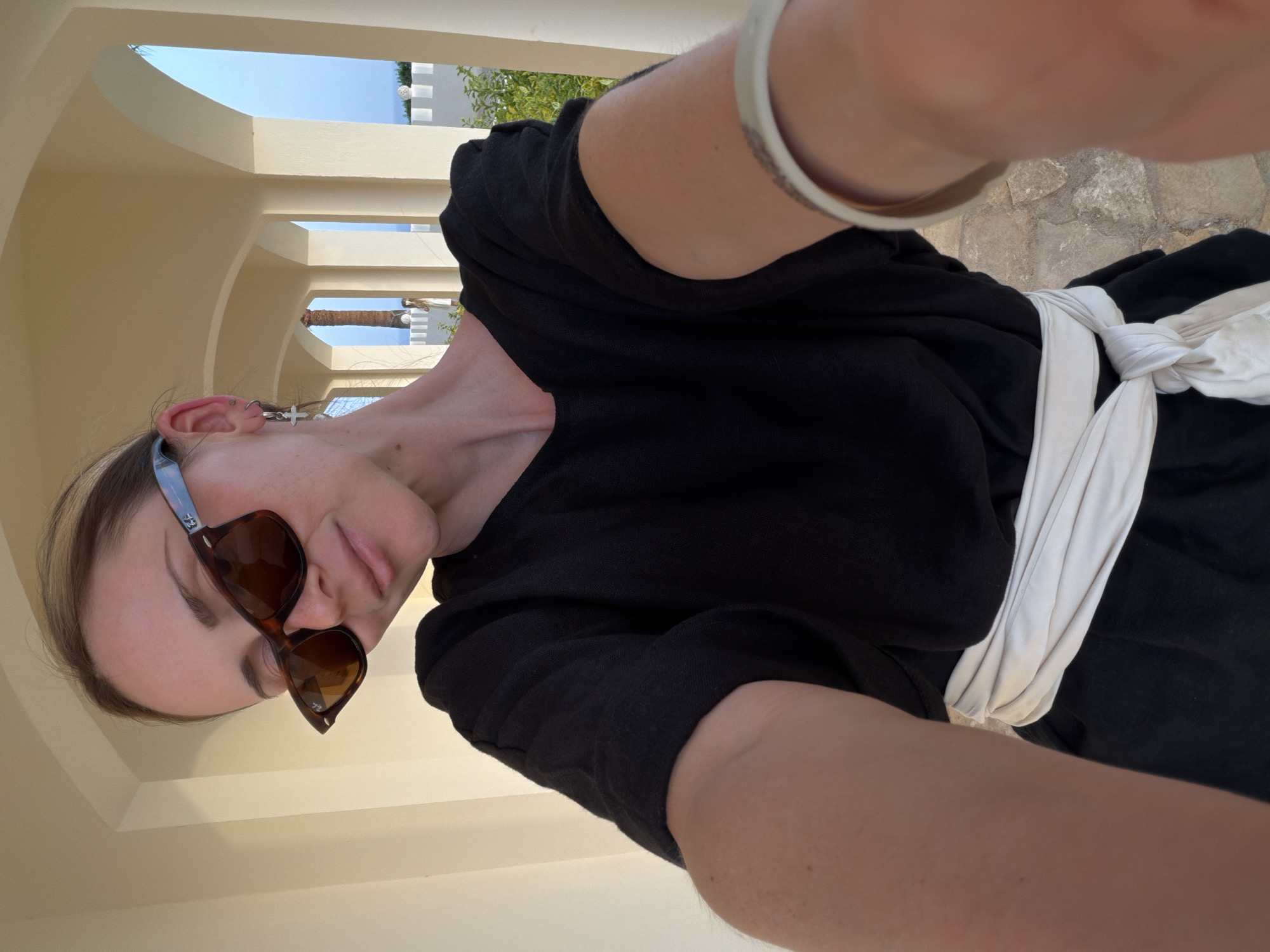 A woman in Sharm El Sheikh, Egypt, takes a selfie. She is wearing sunglasses and a black dress with a white tie. The location is an outdoor corridor with archways. The background shows palm trees and buildings. The flooring is tiled.
