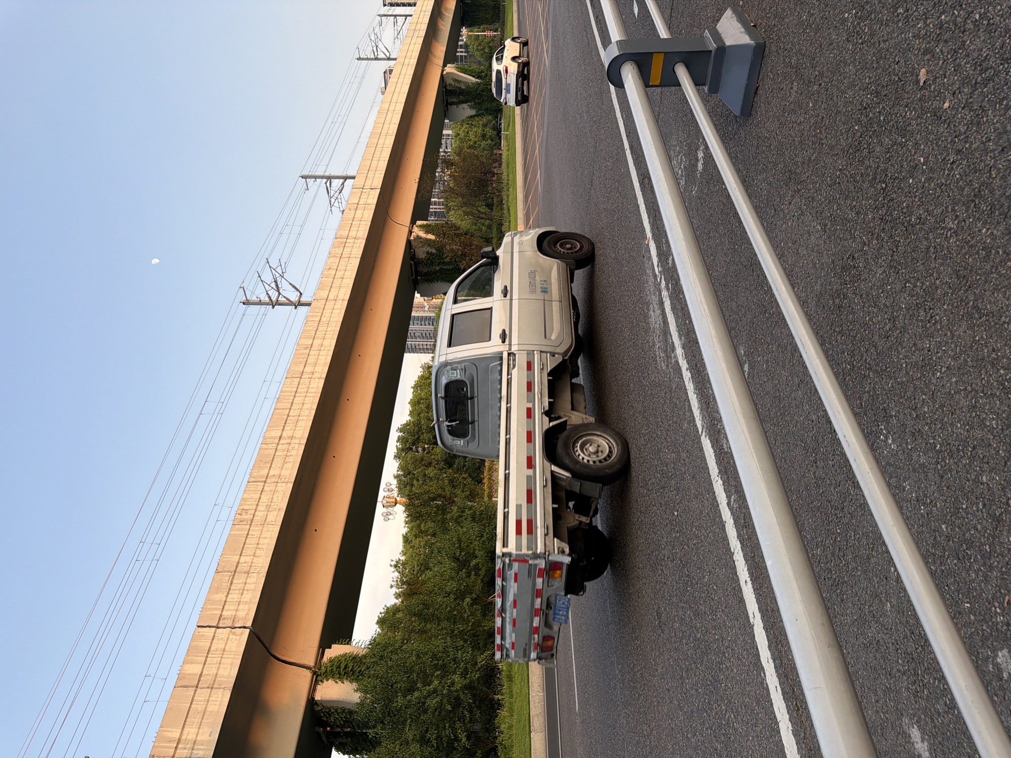 In Jiangjin, China, at a low-angle outdoor shot, a silver flatbed truck is driving on a road. The truck has a grey cab and a silver flatbed with red and white reflective strips. The truck is traveling on a paved road with lane markings. In the background, a raised concrete overpass with overhead power lines runs across the scene. On the left side of the overpass, green ivy climbs up the concrete supports. Beyond the overpass, there are trees and some buildings are visible in the distance. The sky is clear and blue, with a sliver of the moon visible. A white SUV is also visible on the road in the distance, traveling in the same direction as the truck. The foreground is partially occupied by a metal guardrail with grey posts and yellow accents.