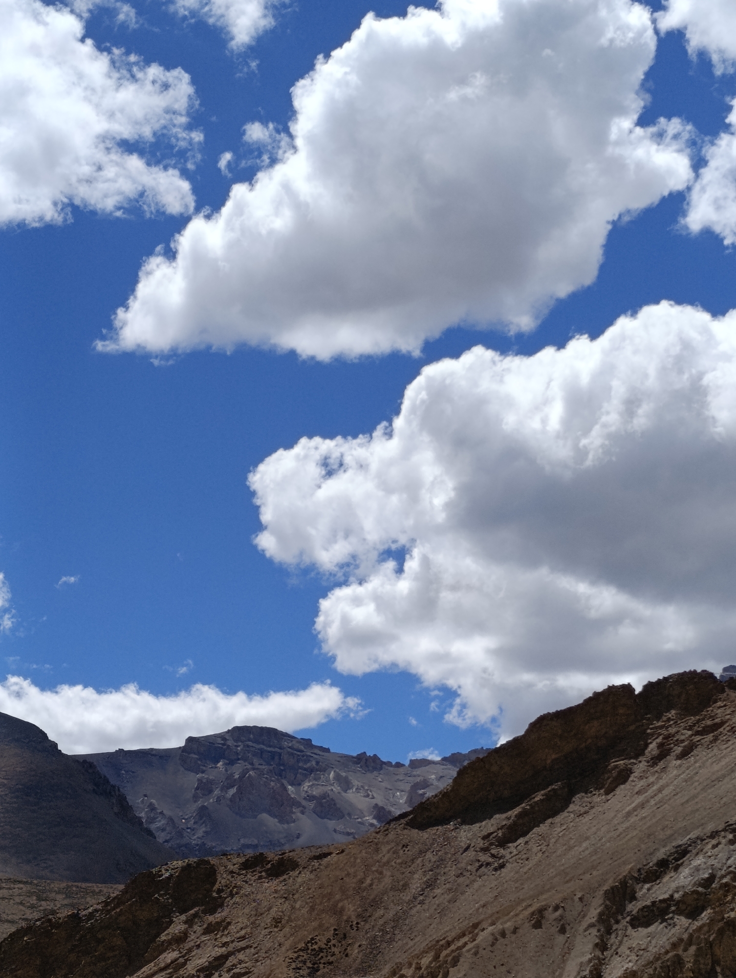 A wide-angle, vertically oriented photograph captures a rugged mountainous landscape under a bright blue sky dotted with fluffy white clouds. The foreground is dominated by a steep, rocky slope covered in loose scree and boulders. The mid-ground features a dramatic rock formation jutting out from the right side of the frame, casting a shadow onto the lower slopes. Further back, towering mountains with jagged peaks rise under a vast expanse of sky. The lighting suggests it is daytime, likely midday, given the intensity of the sunlight and the clarity of the sky. The overall impression is one of remote, arid, and imposing natural beauty. The location is identified as Pang, India, a high-altitude settlement in Ladakh known for its challenging terrain and stunning mountain scenery.